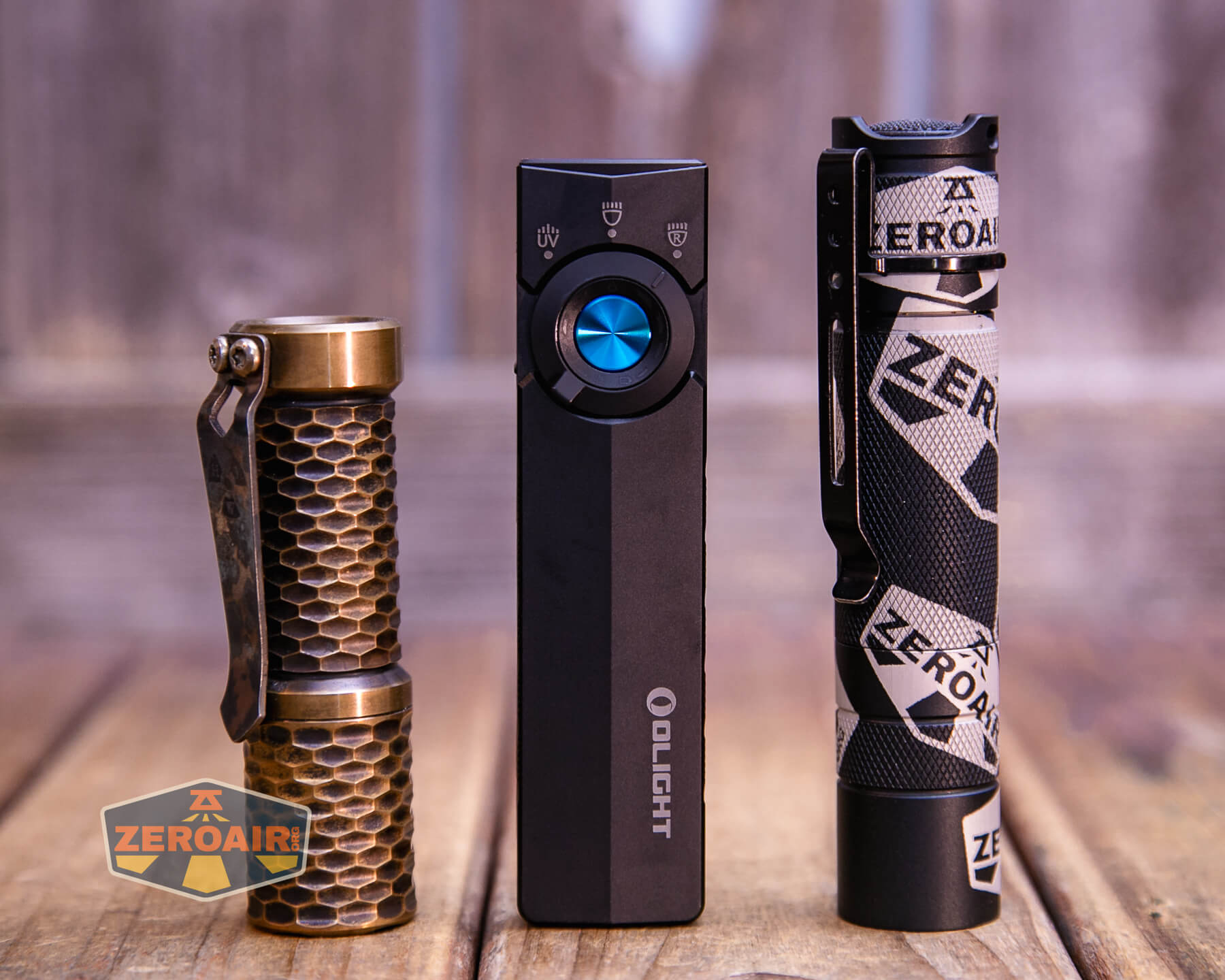 Three flashlights stand upright on a wooden surface: one with a brass honeycomb design, a rectangular black one with a blue button, and a black one with ZEROAIR branding and a patterned clip.