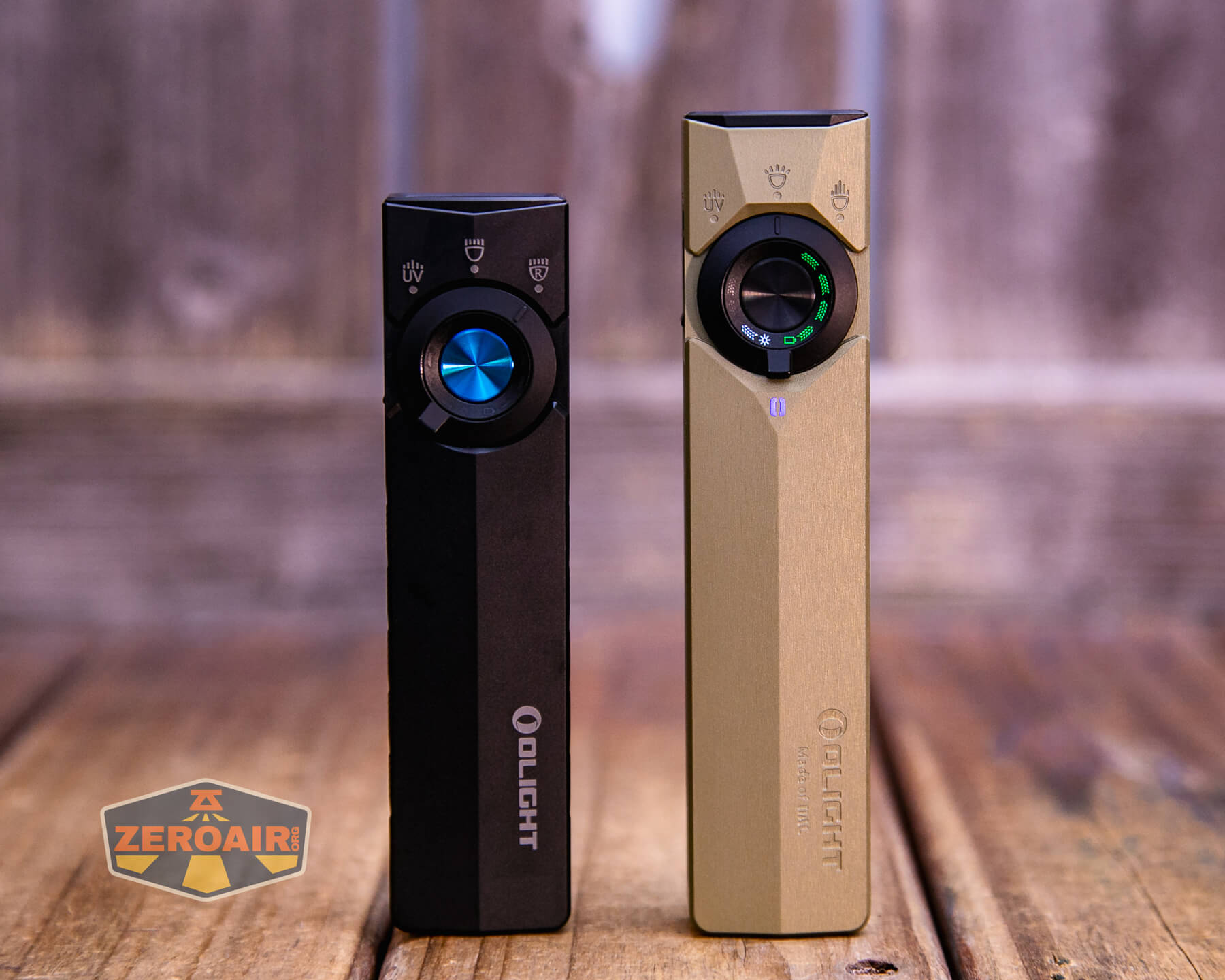 Two rectangular Olight flashlights, one black and one gold, stand upright on a wooden surface. Both have circular blue power buttons and indicator lights. The ZeroAir logo is visible on the lower left.