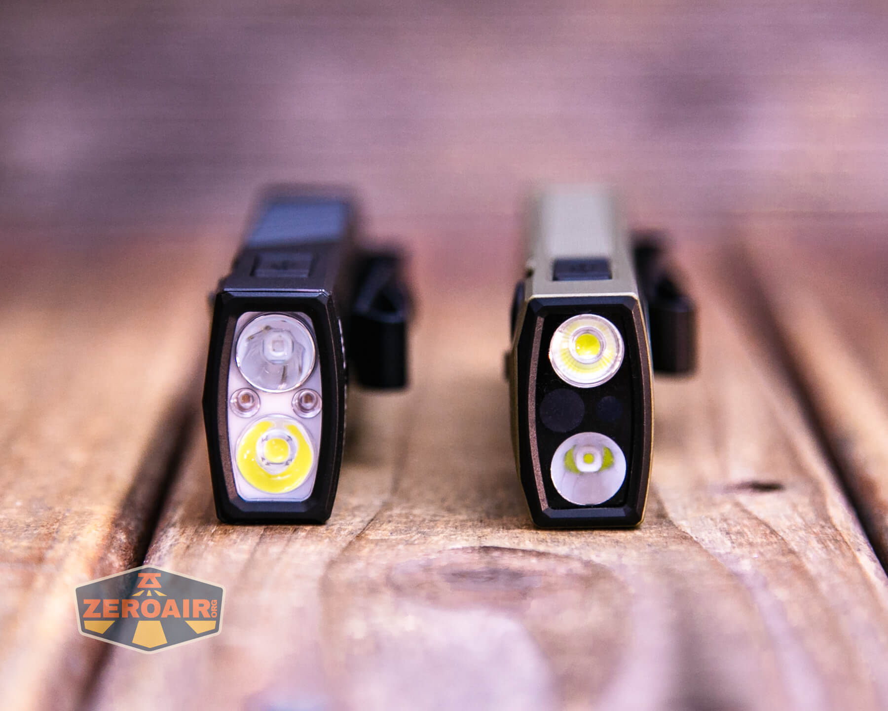 Two rectangular flashlights are standing upright on a wooden surface. Each has three visible LED lights on the front. The flashlight on the left is black; the one on the right is olive green. A ZeroAir logo is in the lower left corner.