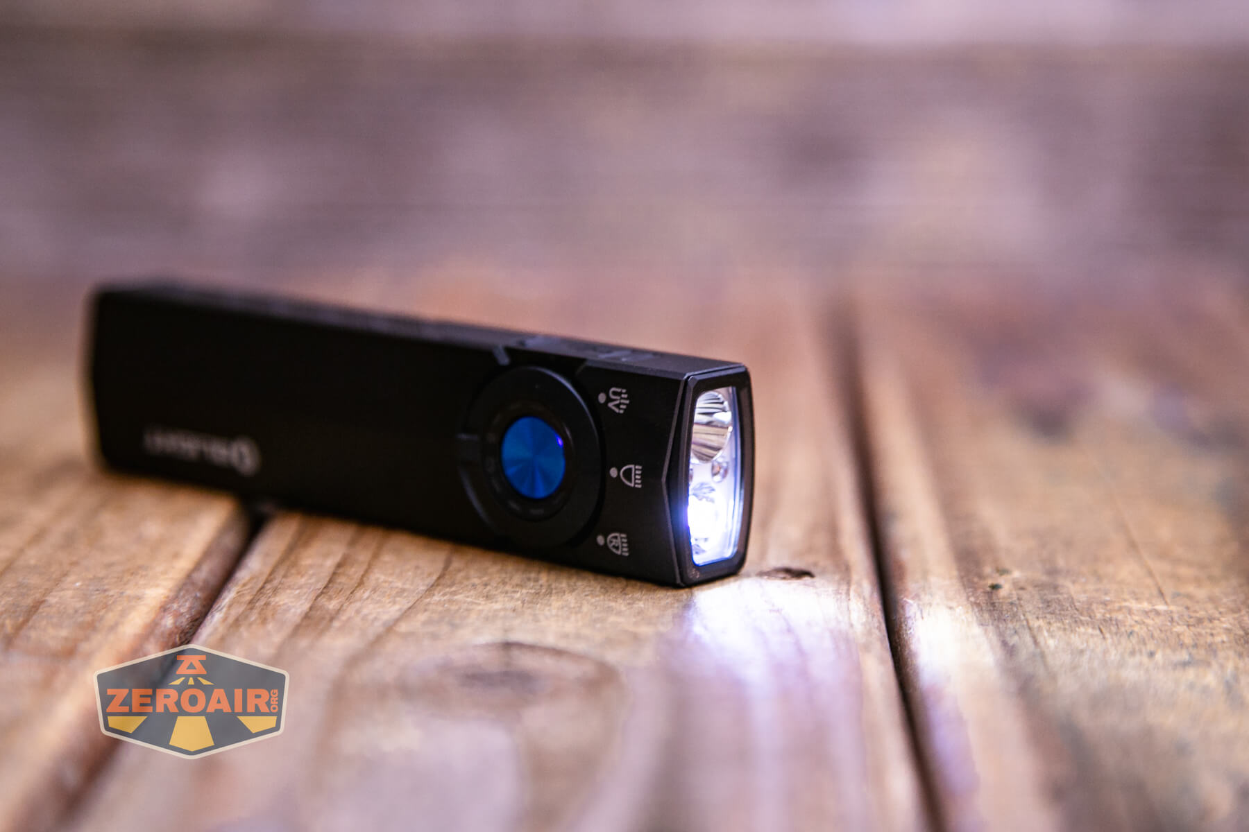 A small black flashlight with a blue button is turned on and rests on a wooden surface. The flashlight beams light forward. A ZeroAir logo is visible in the lower left corner of the image.