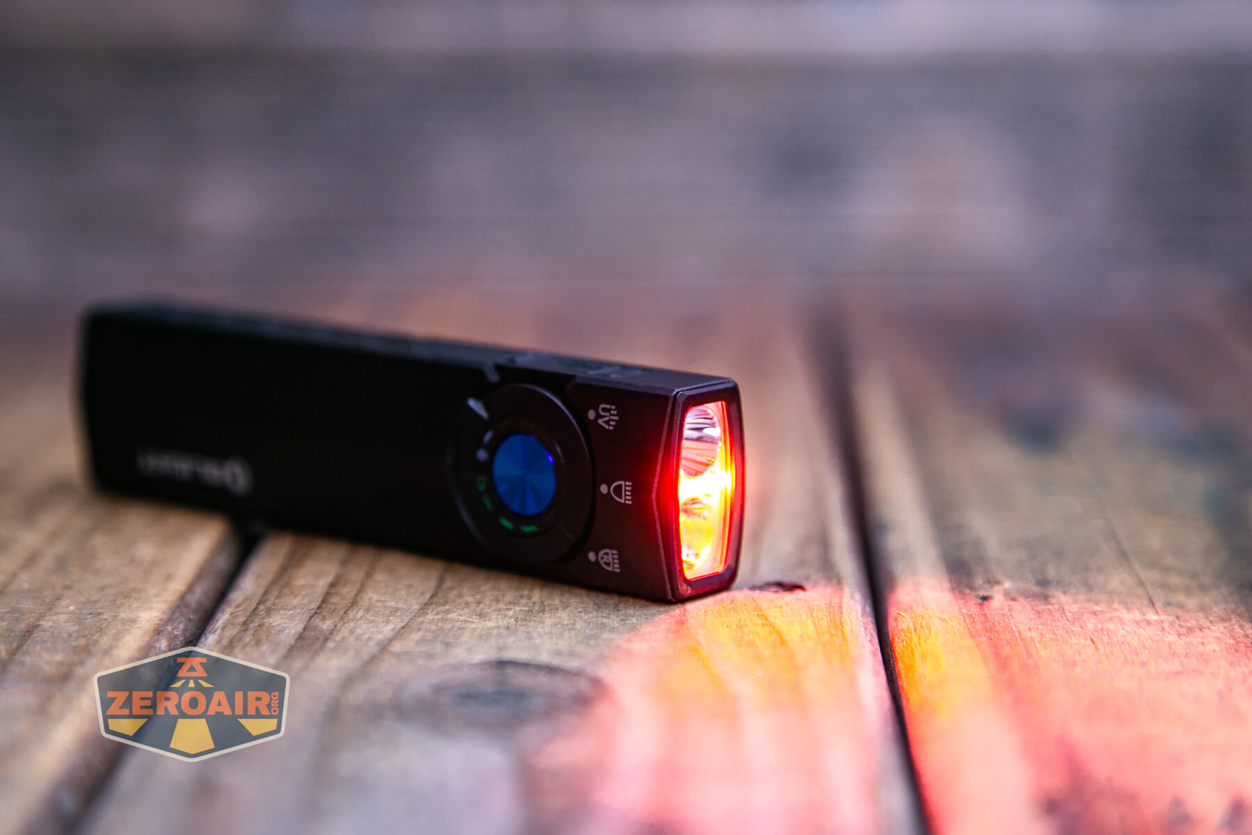 A black Olight flashlight lies on a wooden surface, emitting a red light from its front. The focus is on the illuminated end, and an orange ZeroAir logo is visible on the bottom left corner.
