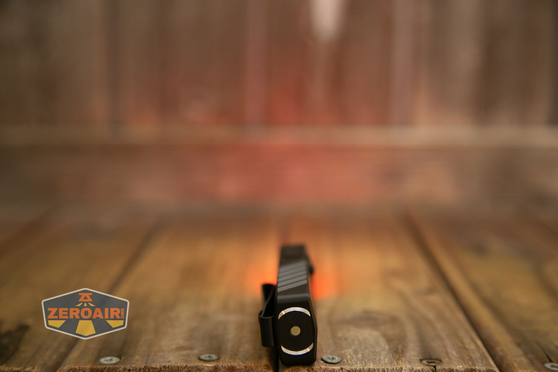 A small black flashlight with a side clip is placed upright on a wooden surface. The background is out of focus with warm reddish lighting. A ZeroAir logo appears in the lower left corner.