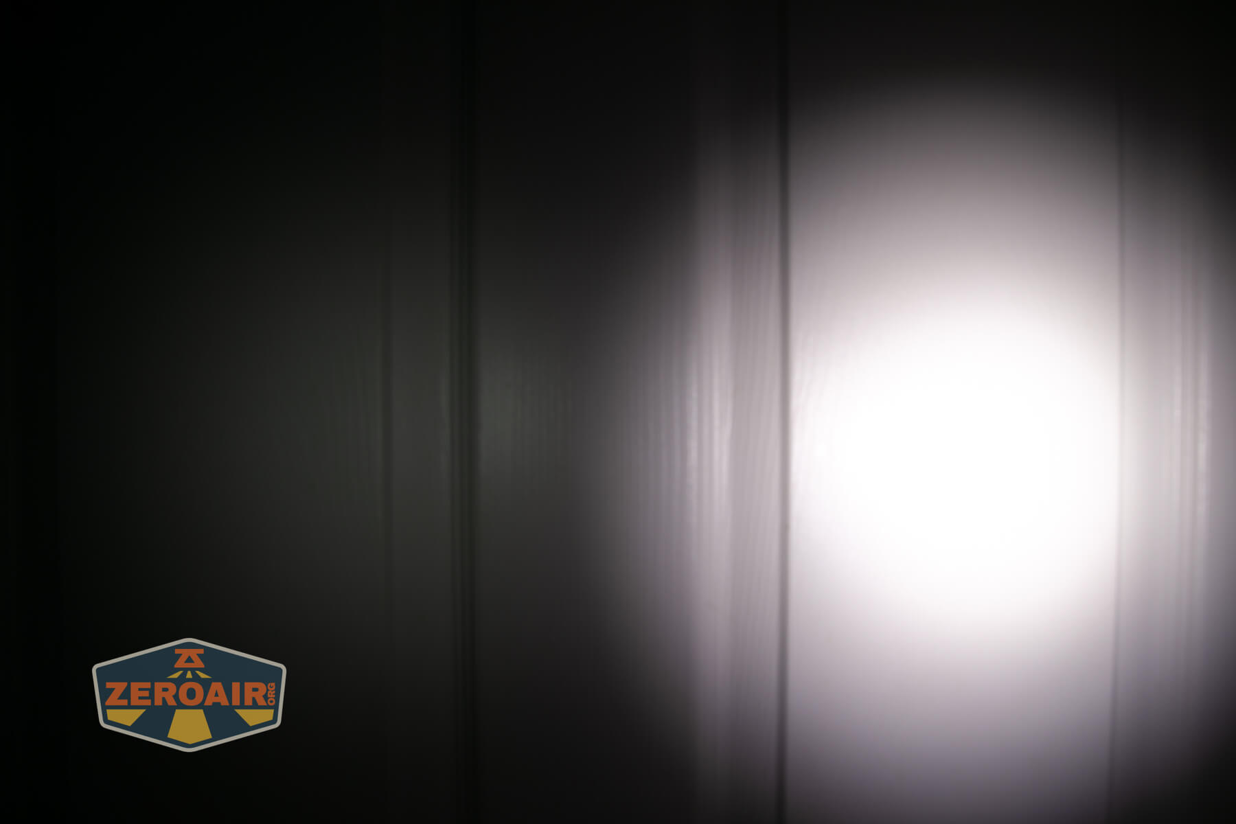 A bright, circular flashlight beam shines on a dark, vertically paneled surface. The light creates visible shadows and texture. A ZEROAIR logo is in the lower left corner of the image.