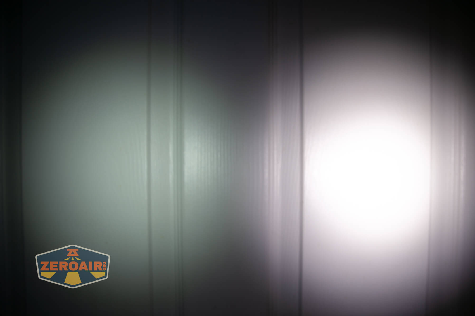 A flashlight beam with a bright white center and a dimmer green-tinted area is projected onto a vertically paneled wall. The ZeroAir logo is visible in the bottom left corner.