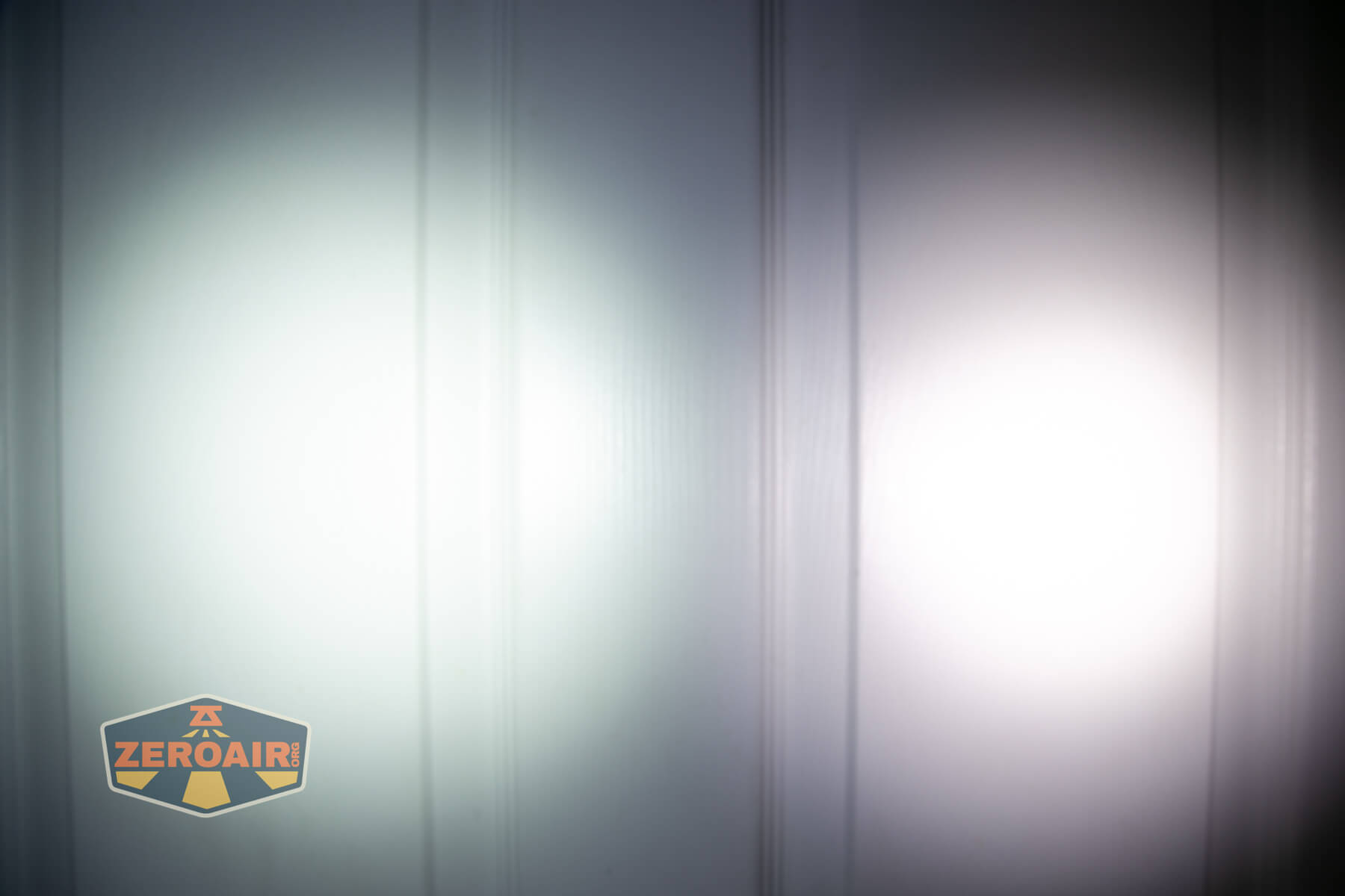 Two circular beams of white light are projected onto a vertically paneled white surface. The logo ZEROAIR appears in the bottom left corner of the image.