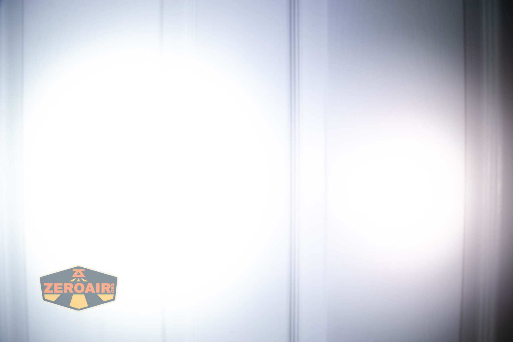 A bright white flashlight beam shines on a white wall with vertical lines. The ZeroAir logo is visible in the lower left corner of the image.