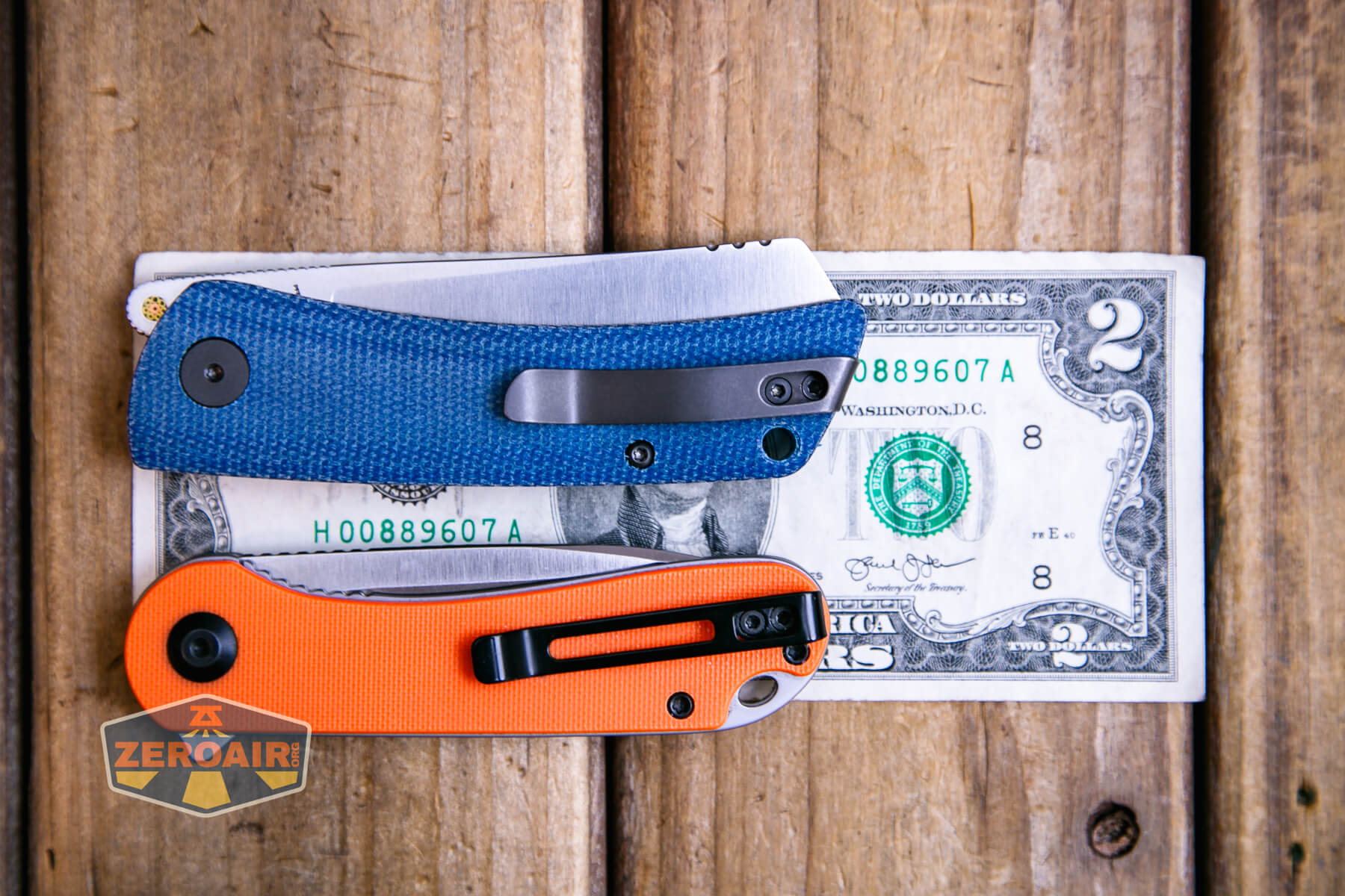 Two folding pocket knives, one with a blue handle and one with an orange handle, are placed closed on top of a two-dollar bill on a wooden surface. A small Zeroair logo patch is visible on the left.