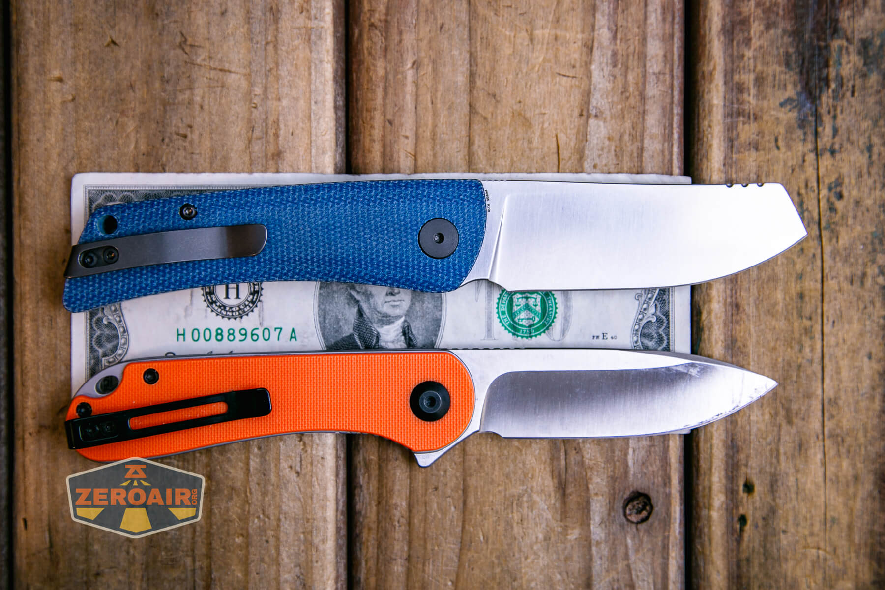 Two folding pocket knives, one with a blue handle and one with an orange handle, are placed on top of a US two-dollar bill on a wooden surface. A Zeroair sticker is also visible in the lower left corner.