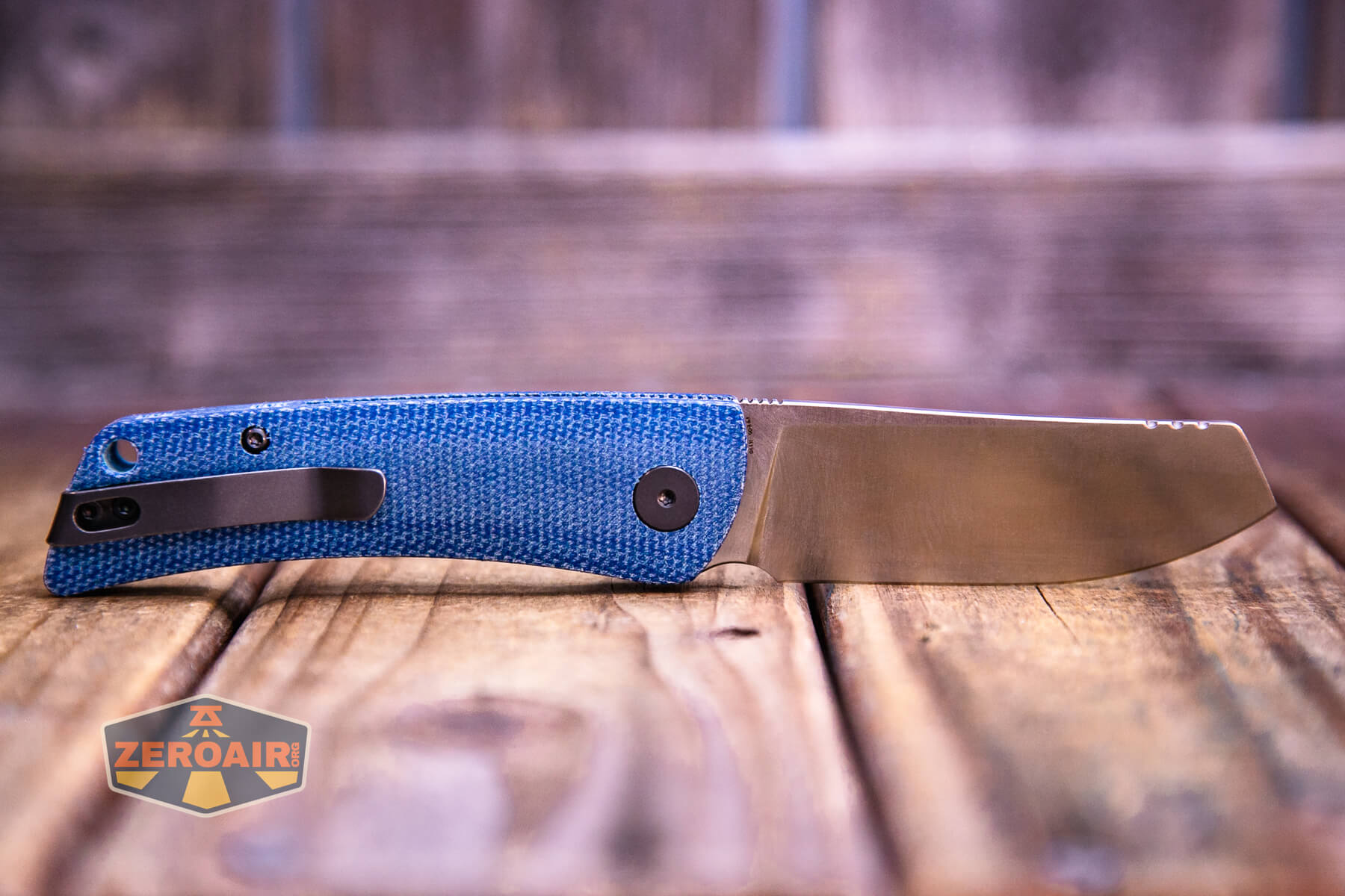 A folding knife with a blue textured handle and a silver blade is displayed on a wooden surface. The knife is partially open, and a ZeroAir logo is visible in the bottom left corner.