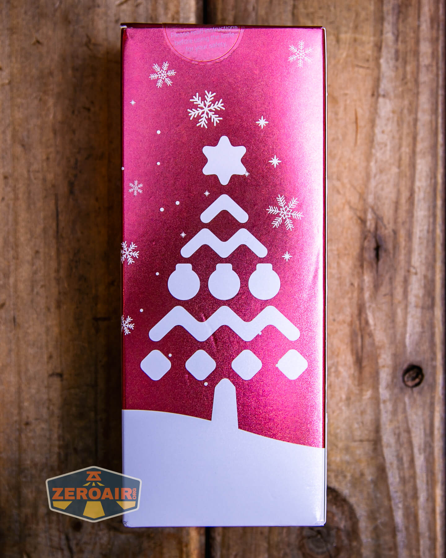 A rectangular pink box with a white stylized Christmas tree and snowflakes design is set on a wooden surface. A logo with the text ZEROAIR appears in the lower left corner.