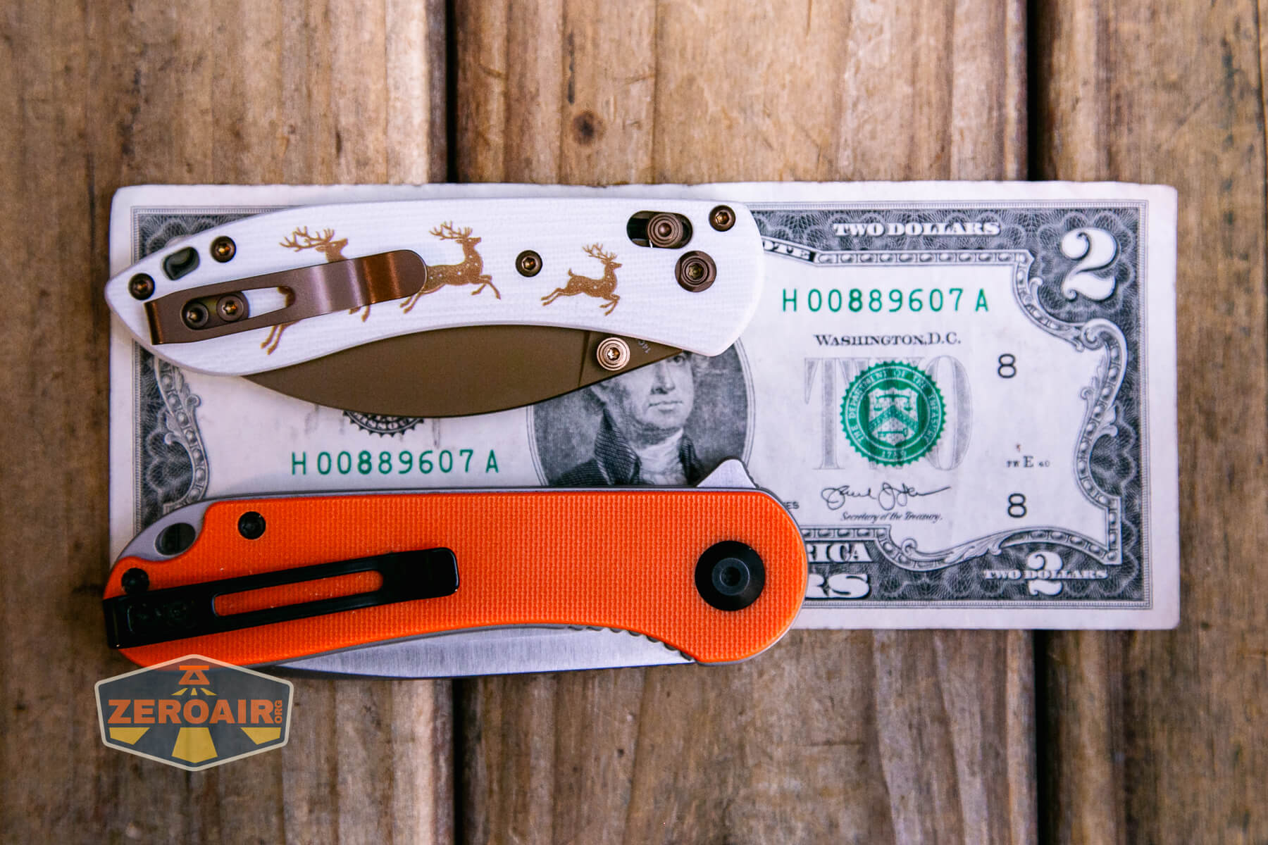 Two folding pocket knives, one with an orange handle and one with a white handle featuring reindeer designs, are placed on top of a US two-dollar bill on a wooden surface. A ZEROAIR logo is visible in the corner.
