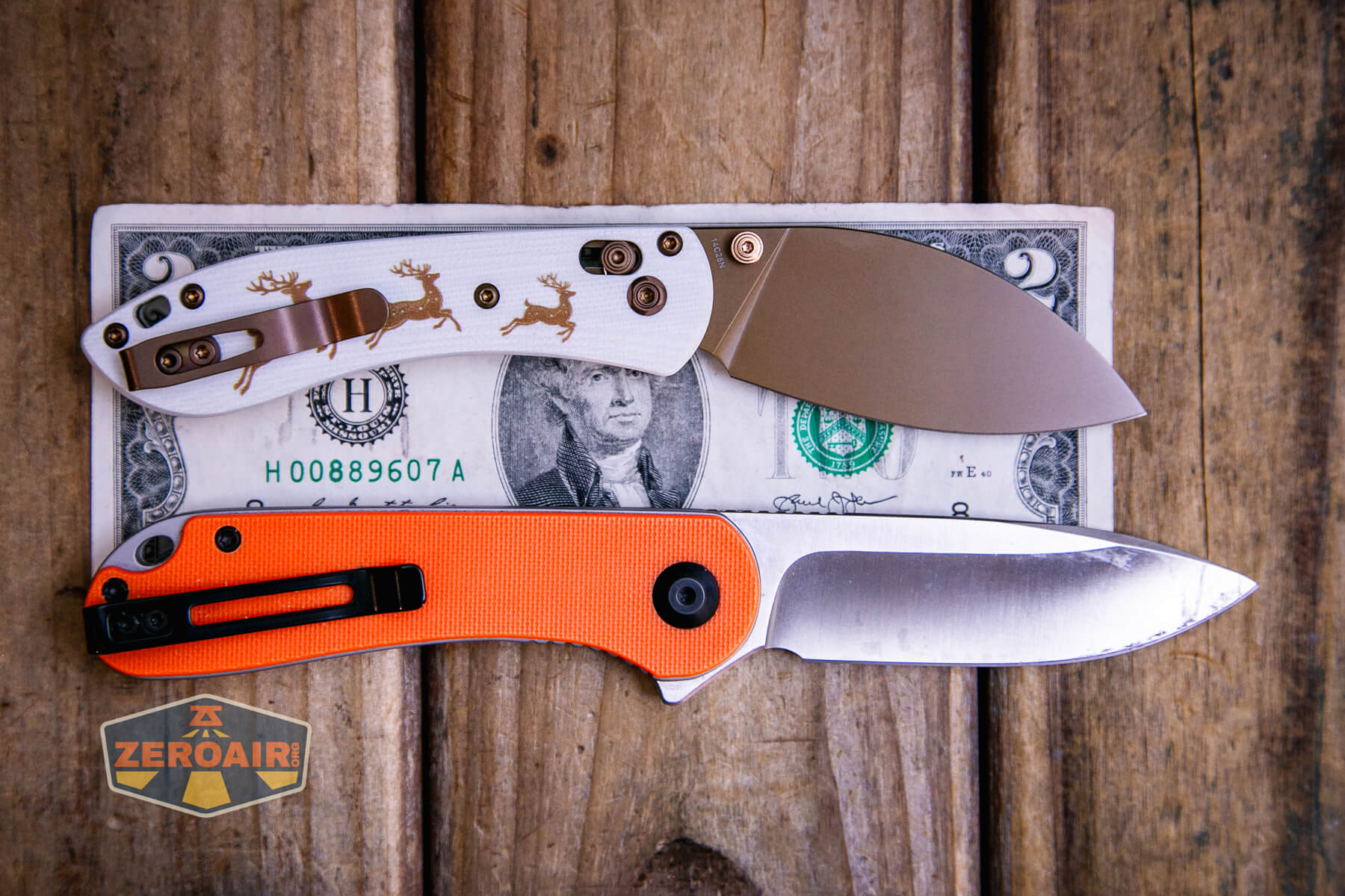 Two folding knives, one with a white handle decorated with moose and one with an orange handle, are displayed on top of a U.S. two-dollar bill on a wooden surface. A ZeroAir sticker is visible in the corner.