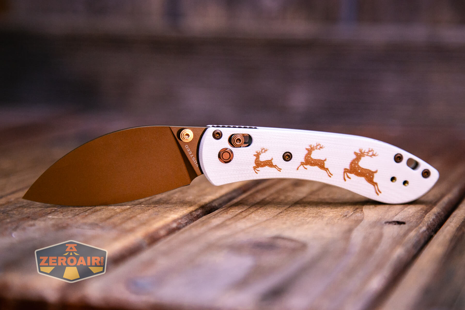 A folding pocket knife with a brown blade and a white handle featuring three brown deer silhouettes, shown on a wooden surface. A ZeroAir logo appears in the lower left corner.