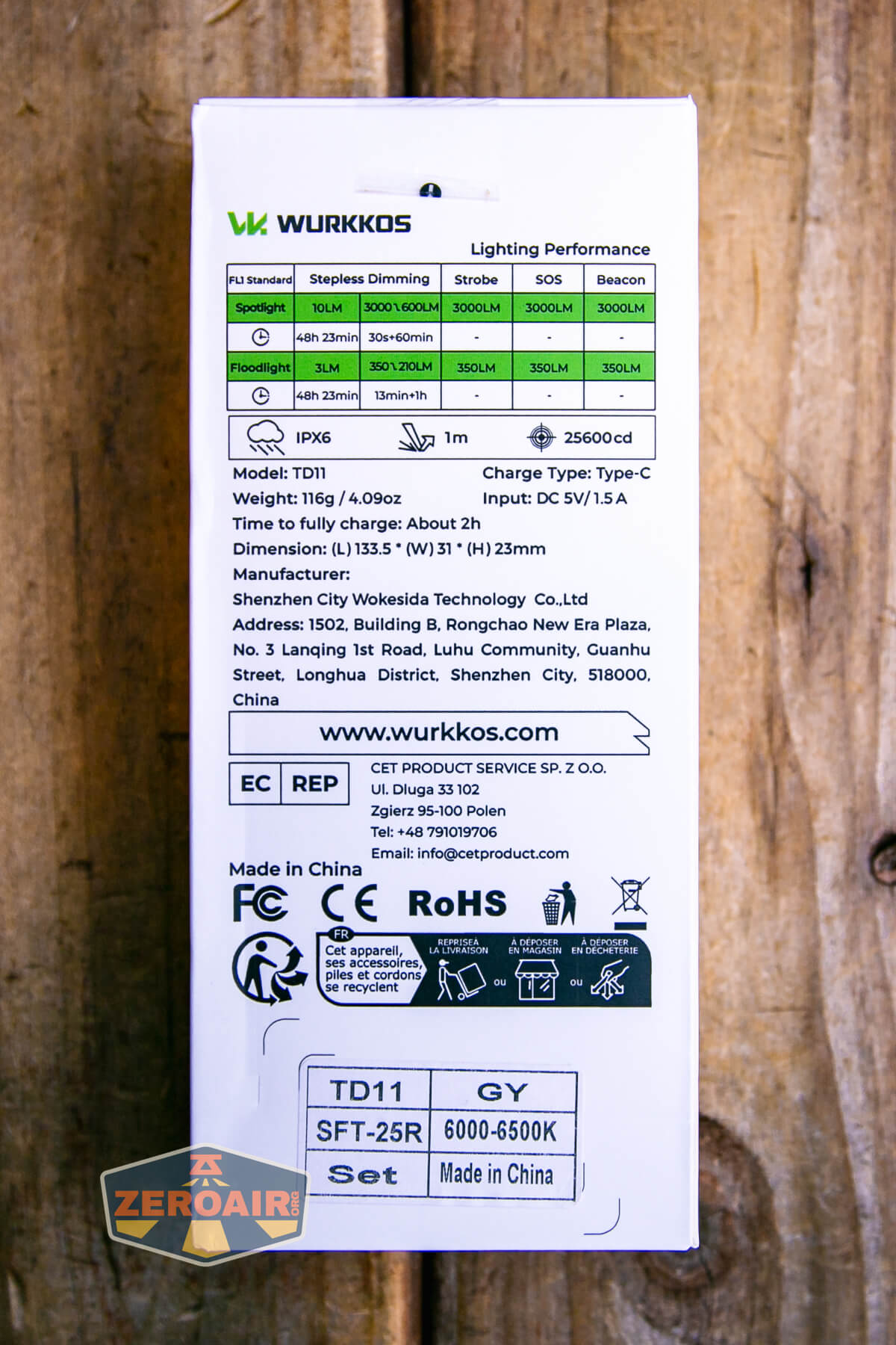 The back of a Wurkkos flashlight box displays technical specifications, lighting performance charts, icons for certifications, company contact details, and model information, all set on a wood background.