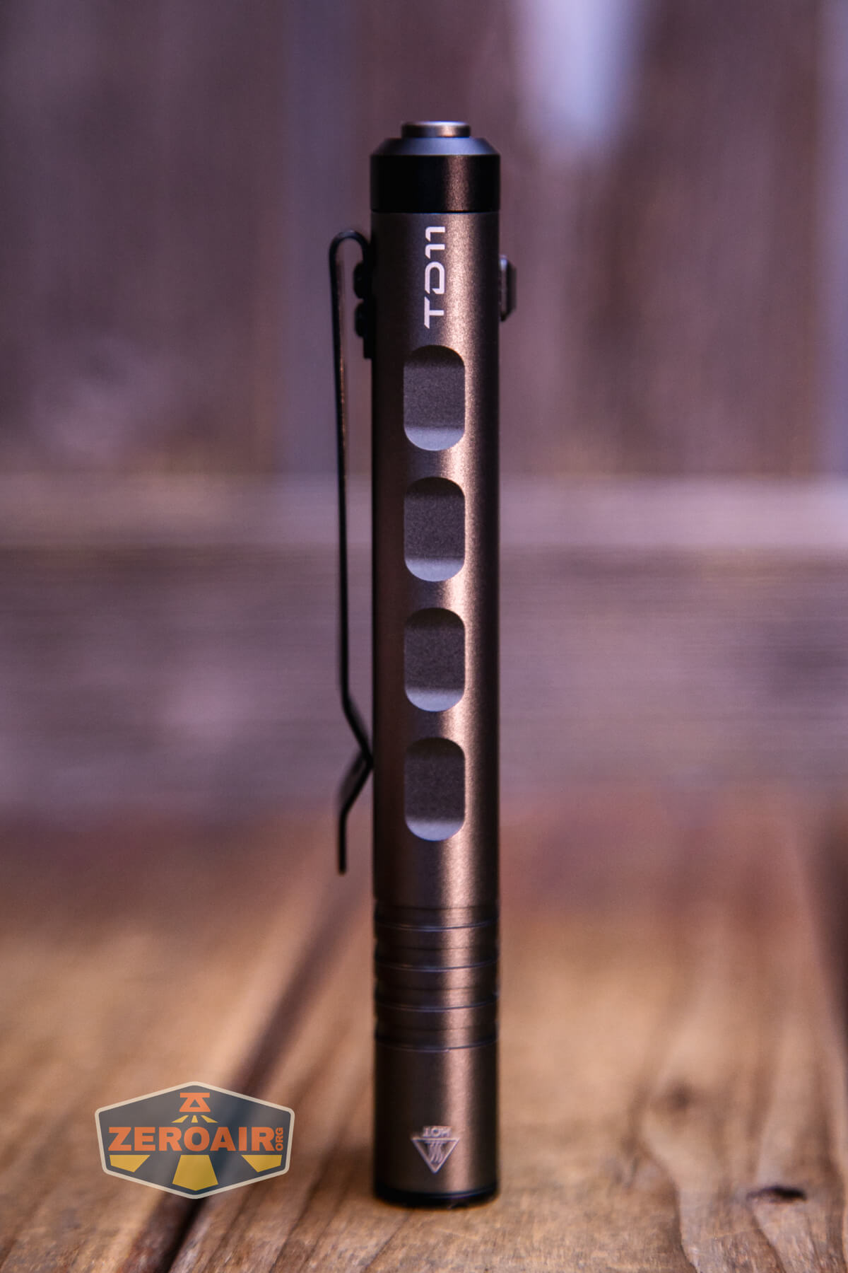 A slim, metallic flashlight with four rectangular cutouts, a side clip, and a button on top stands upright on a wooden surface. A ZeroAir logo is visible in the lower left corner.