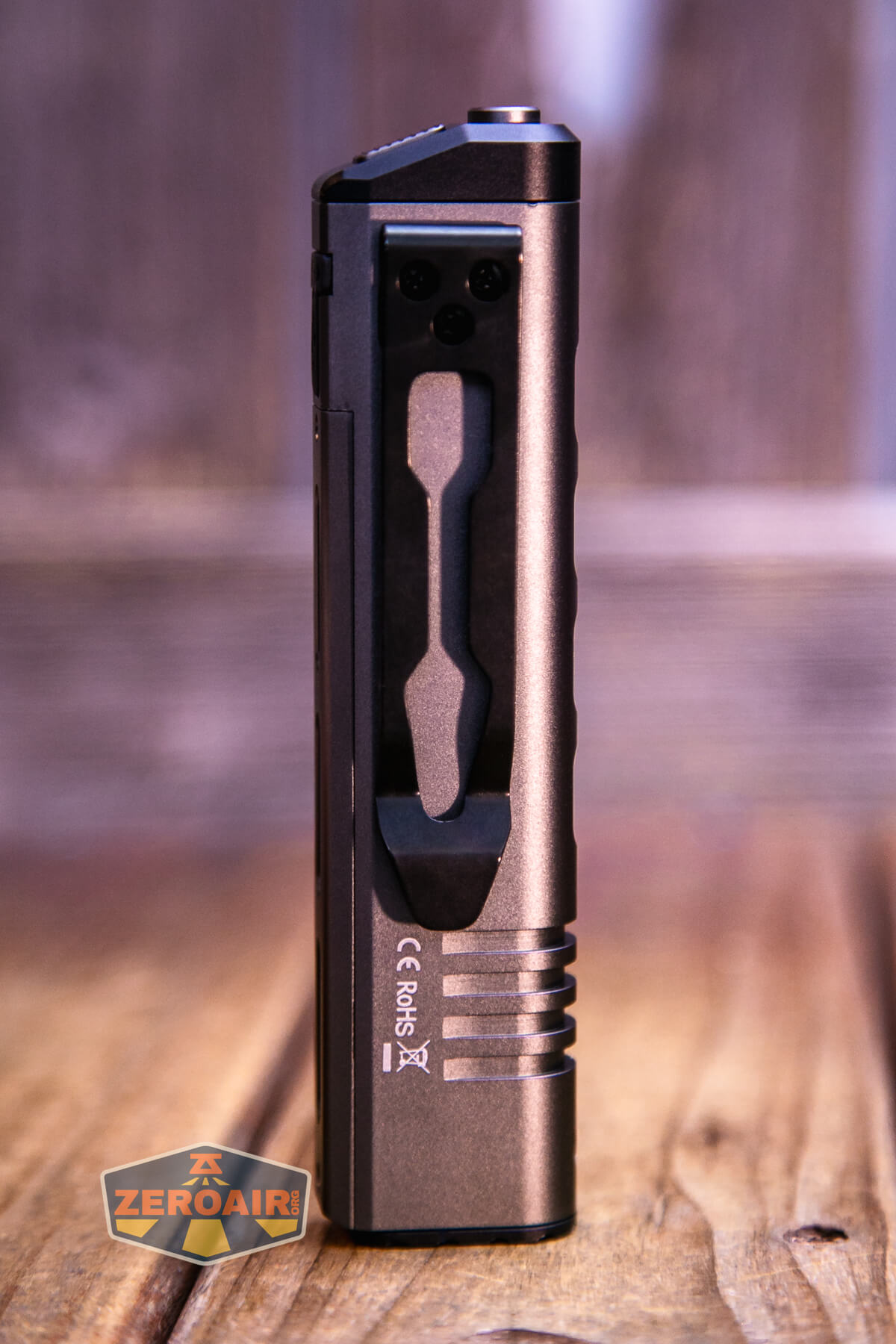 A metallic, rectangular flashlight stands vertically on a wooden surface, showing its side with a pocket clip and grooves. The Zeroair logo is in the bottom left corner of the image.