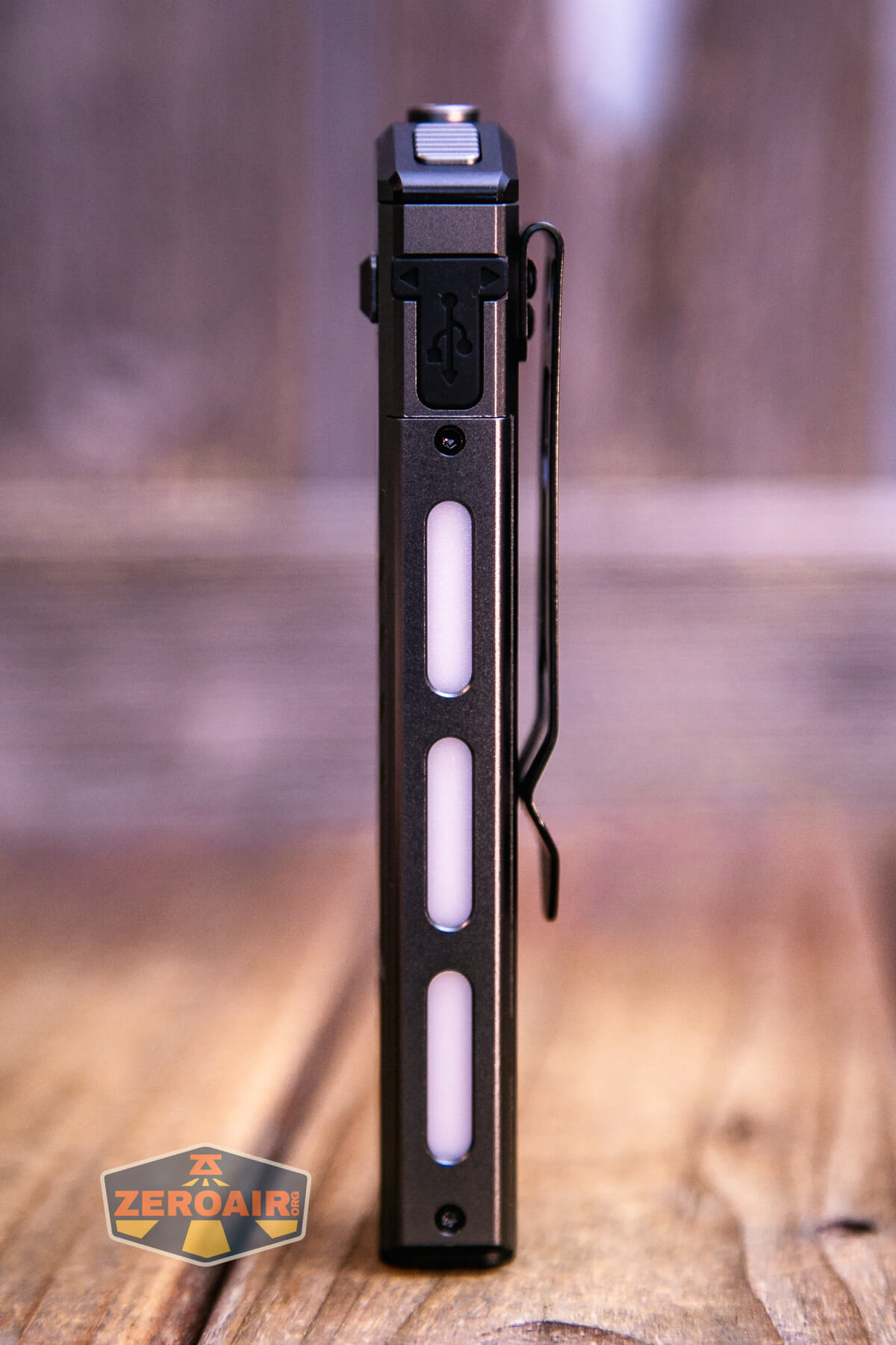 A rectangular, black flashlight with three vertical white light strips and a metal pocket clip is standing upright on a wooden surface. The ZeroAir logo appears in the lower left corner.