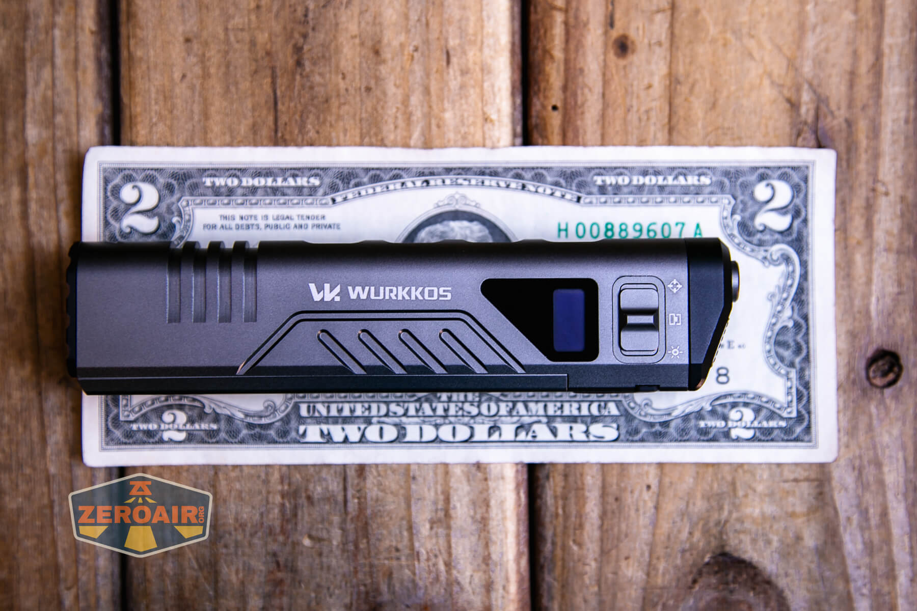 A Wurkkos branded flashlight is placed on top of a two-dollar bill, both lying on a wooden surface. A Zeroair logo is visible in the lower left corner of the image.