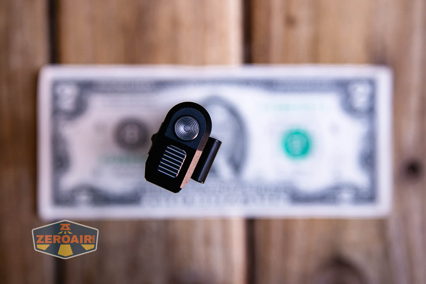 A small black and silver flashlight viewed from above, positioned on top of a blurred U.S. one dollar bill, with a ZeroAir logo in the bottom left corner. The background is wooden.