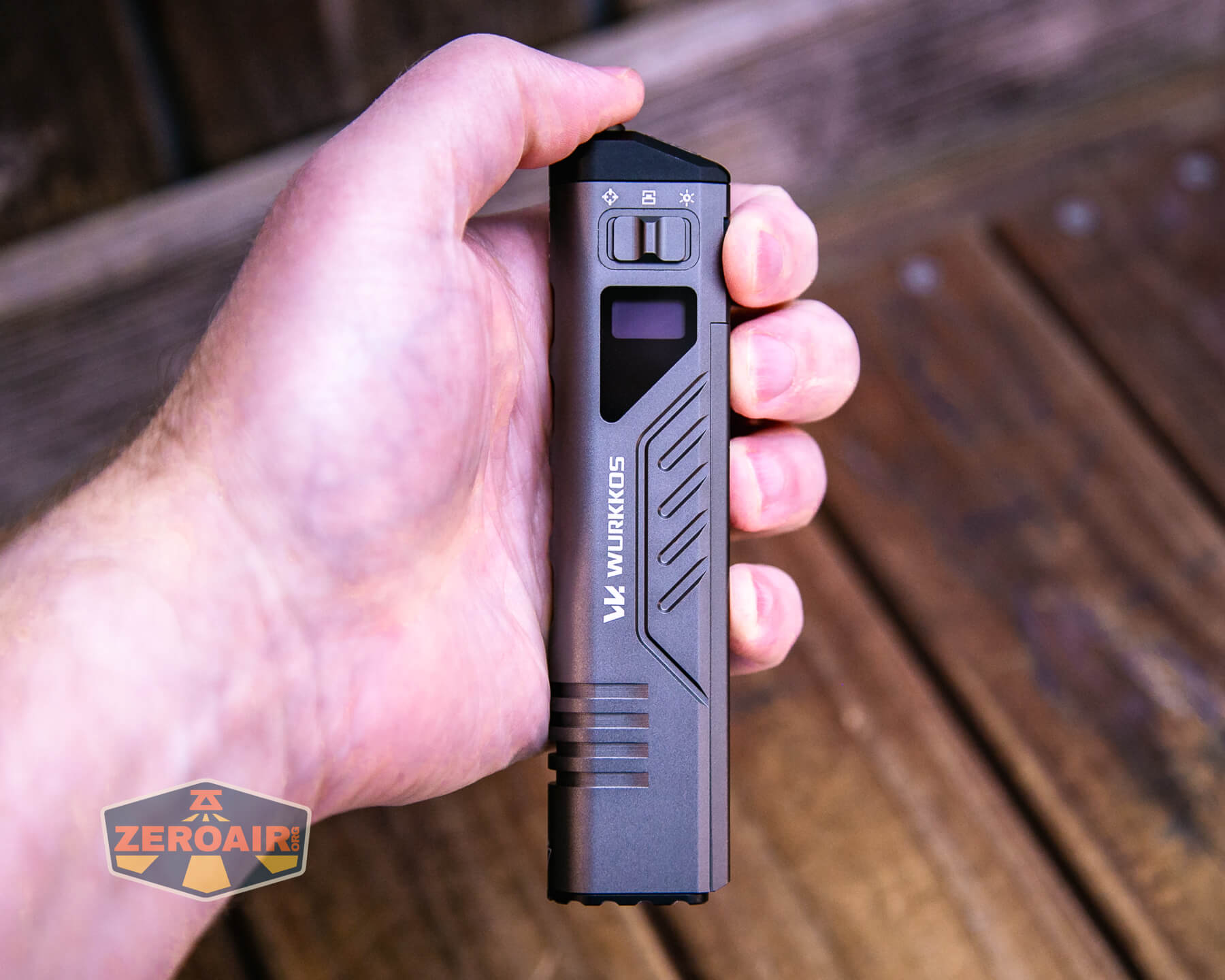 A hand holds a black Wurkkos branded flashlight against a wooden background. The flashlight has visible buttons and indicator icons near the top.