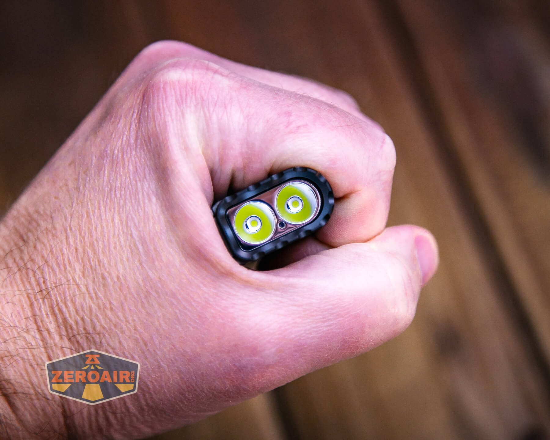 A hand grips a small flashlight, showing two LED bulbs at the front. The background is out of focus, and a ZEROAIR logo is visible in the bottom left corner.
