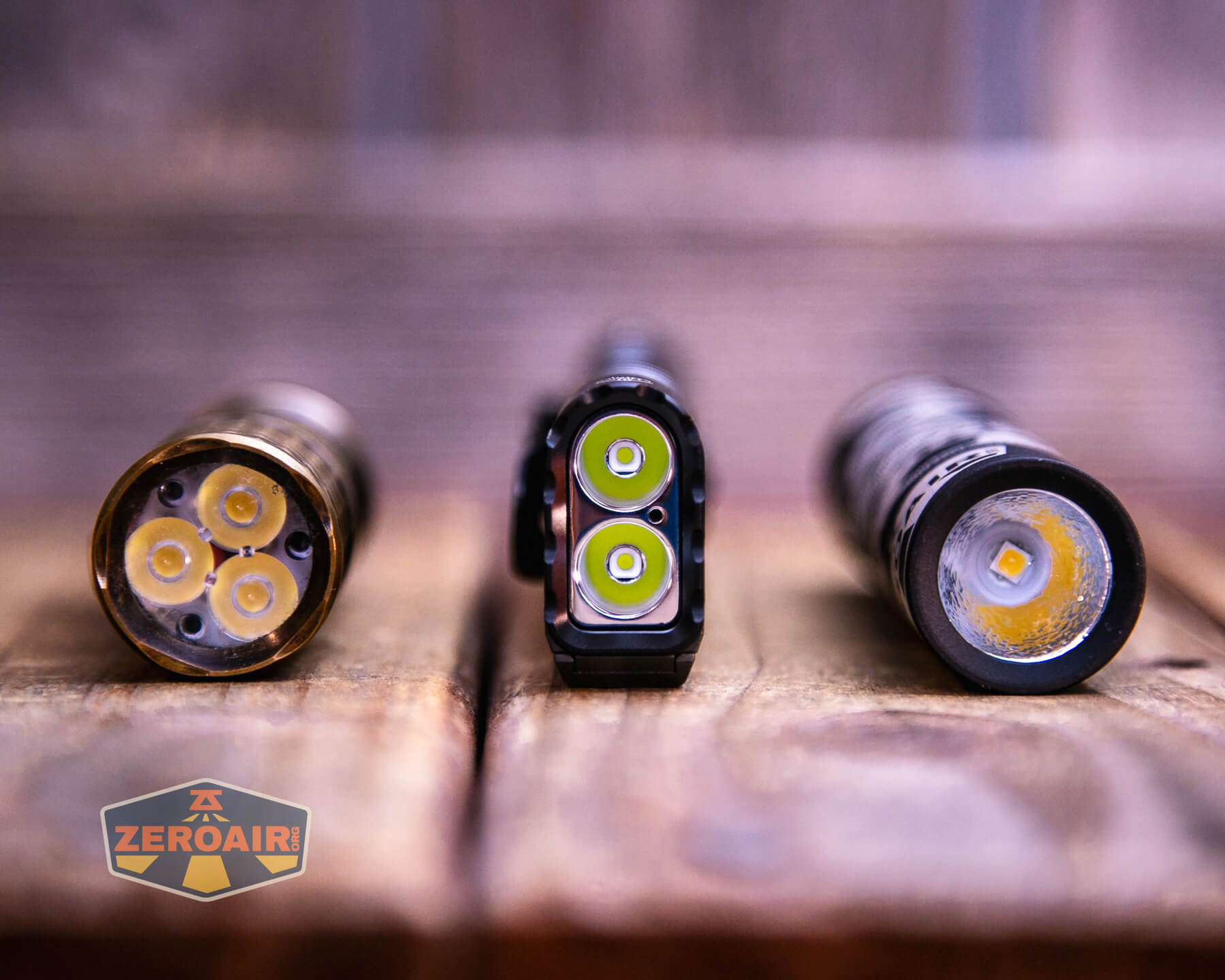 Three flashlights are standing upright on a wooden surface, each showing its LED emitters. The flashlight on the left has four LEDs, the middle has two, and the right has one. A ZEROAIR logo is visible in the lower left corner.