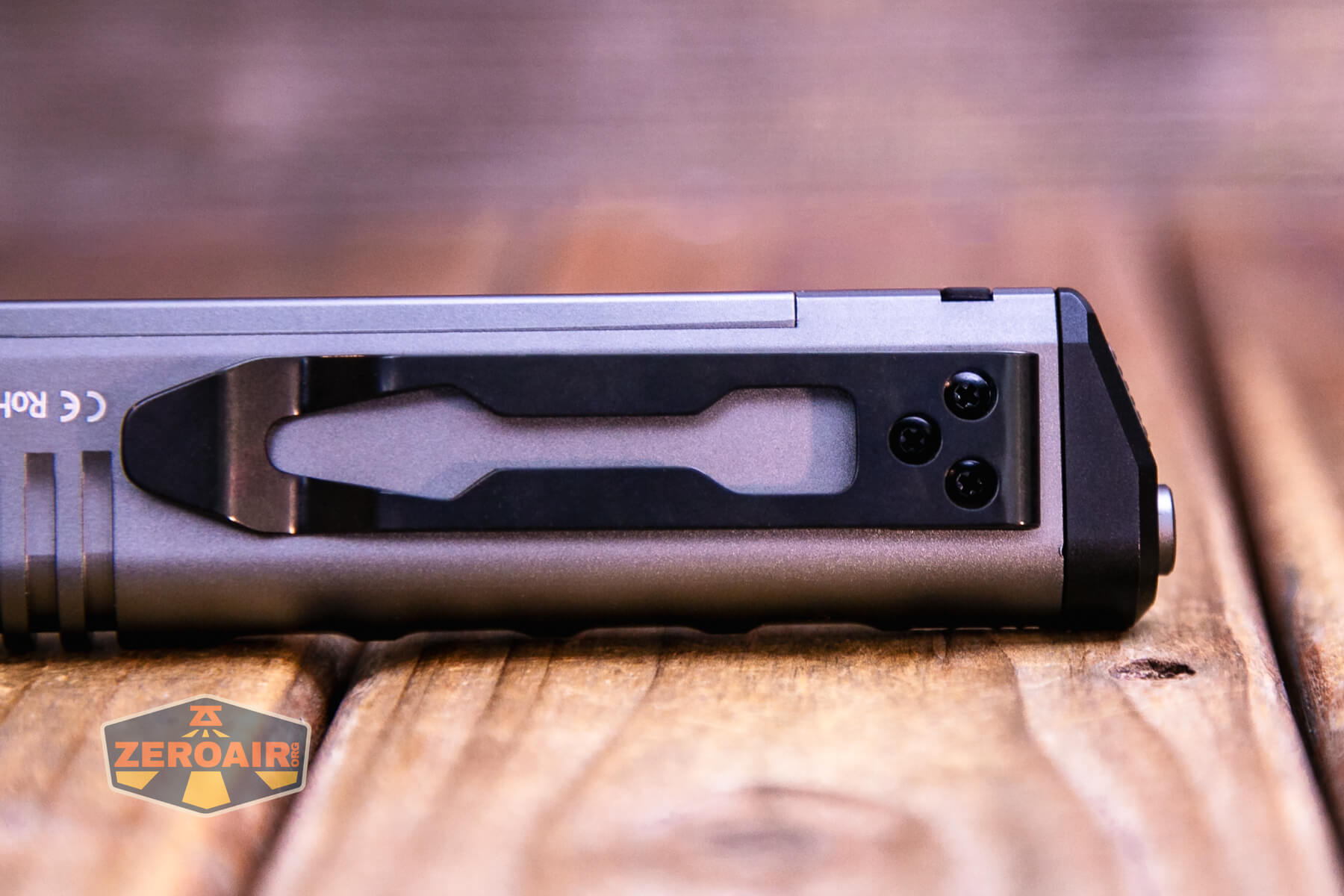 Close-up of a metallic object with a black clip and three visible screws on a wooden surface. The object is angled horizontally, and the background is blurred. A ZeroAir logo is in the lower left corner.