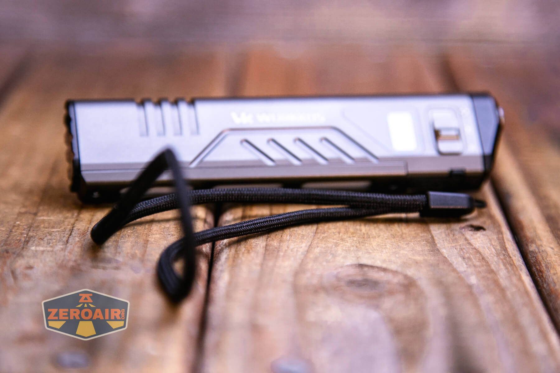 A rectangular flashlight with a wrist strap rests on a wooden surface. The brand name and logo are partially visible on the flashlight, and a ZEROAIR watermark is in the bottom left corner.