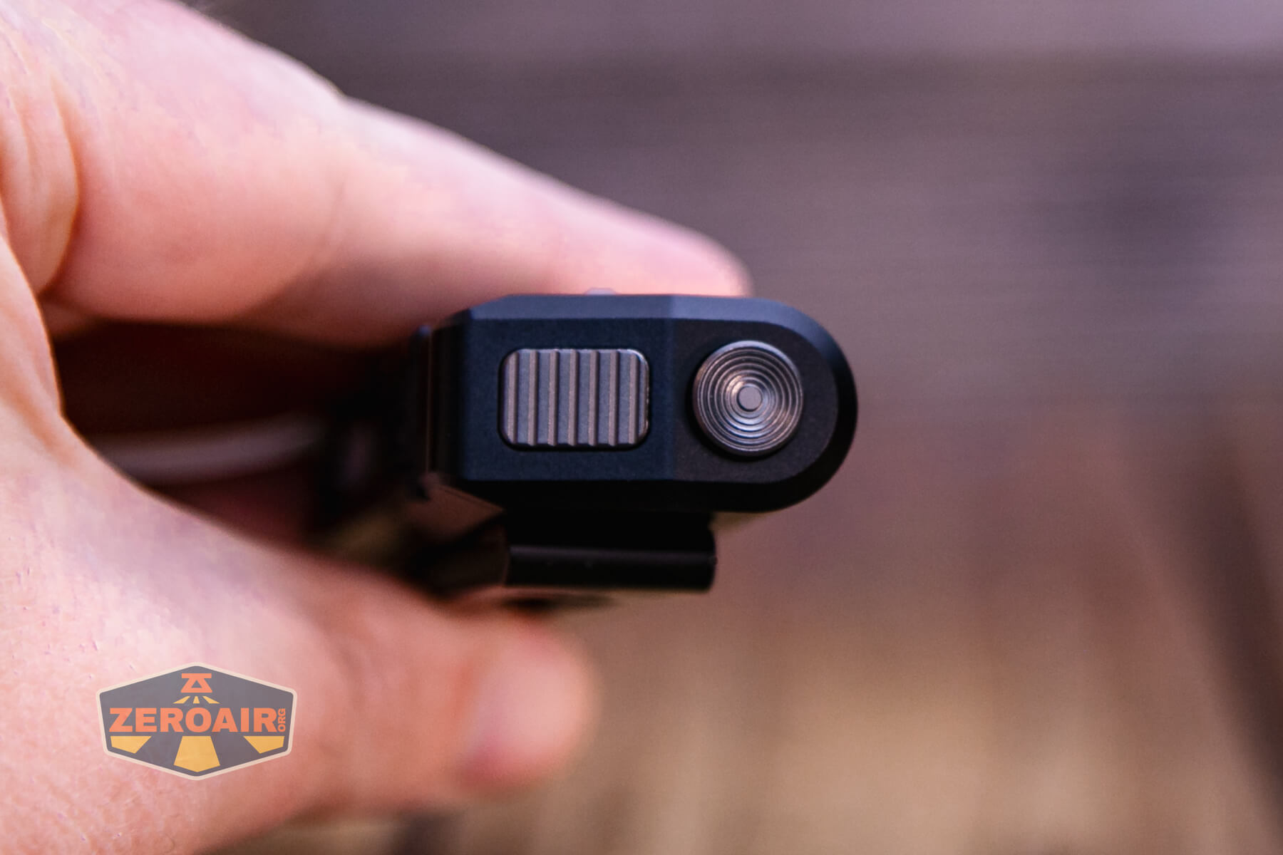 A close-up of a persons hand holding a black rectangular device with a ridged button and a circular metal component. The background is blurred, and a ZeroAir logo is visible in the lower left corner.