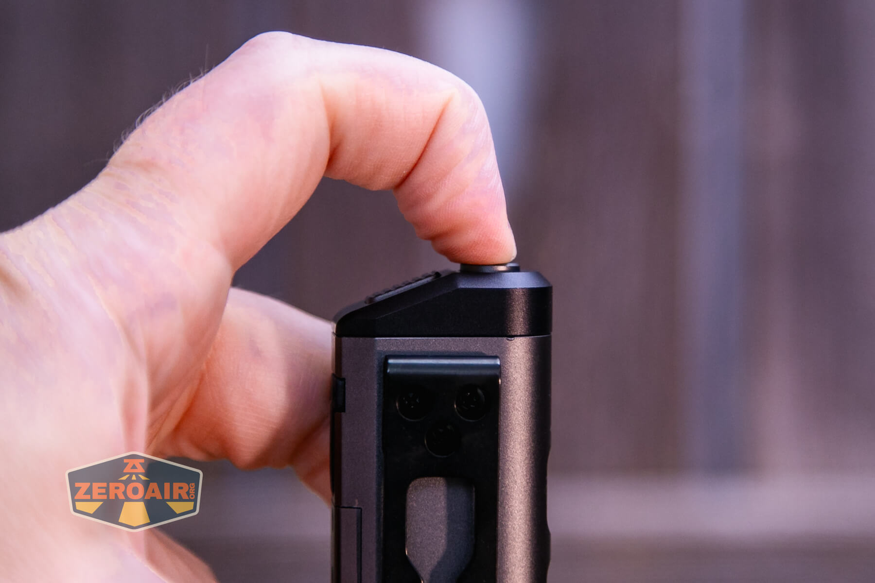 A close-up of a hand pressing a rectangular button on a black electronic device, with a blurred background and a ZeroAir logo in the bottom left corner.