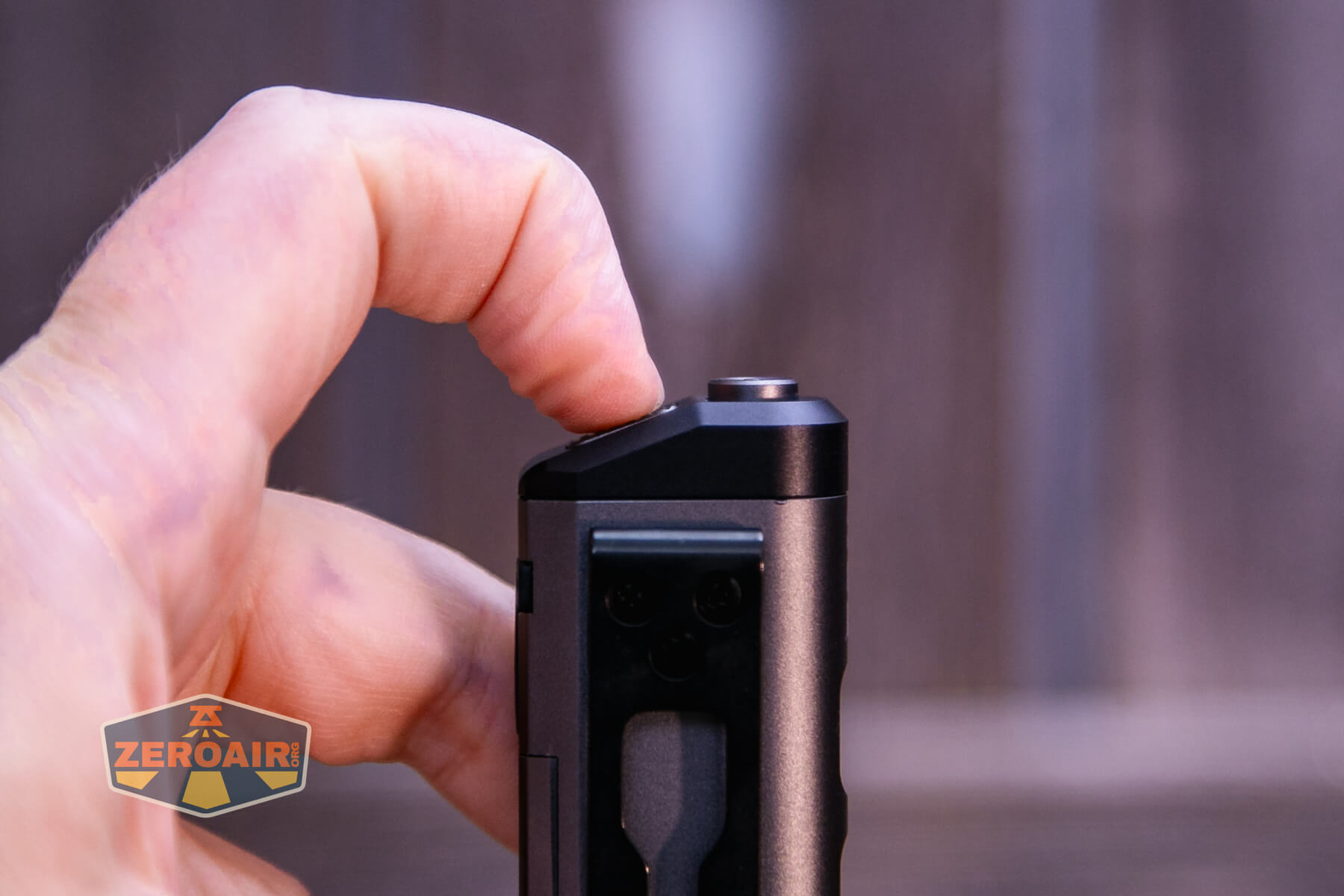 A hand is holding a small black device vertically with a thumb pressing a button on top. The background is blurred, and a ZeroAir logo is visible in the lower left corner.