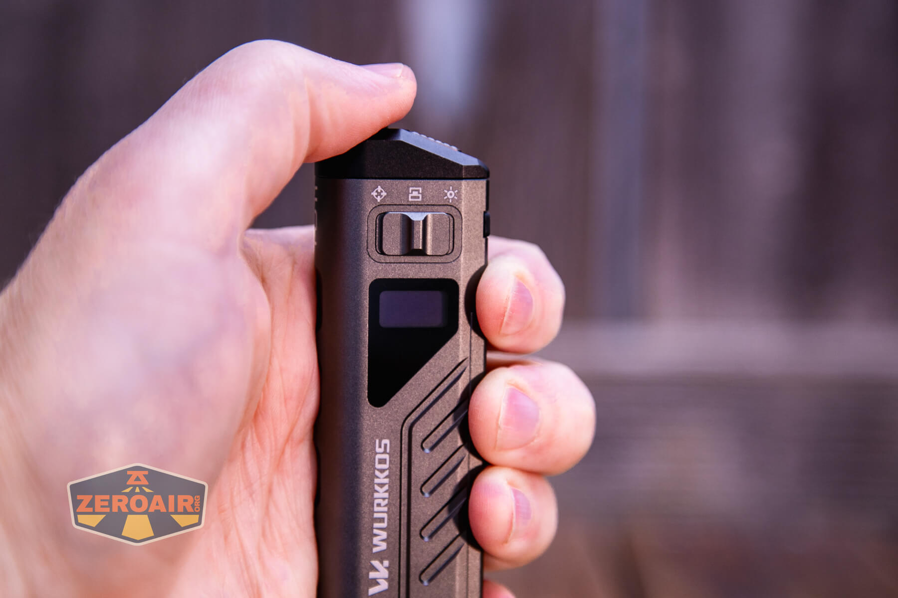 A hand holding a rectangular flashlight with chevron patterns, a digital display, and a switch on the body. The brand Wurkkos is visible on the side. The background is blurred.