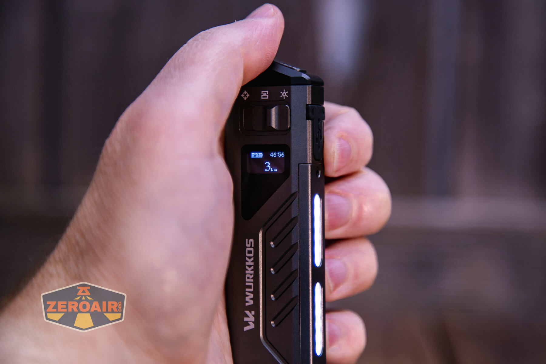 A hand holds a black Wurkkos V4 flashlight, showing its side display screen and illuminated light strips. The display shows battery information. The background is blurred.