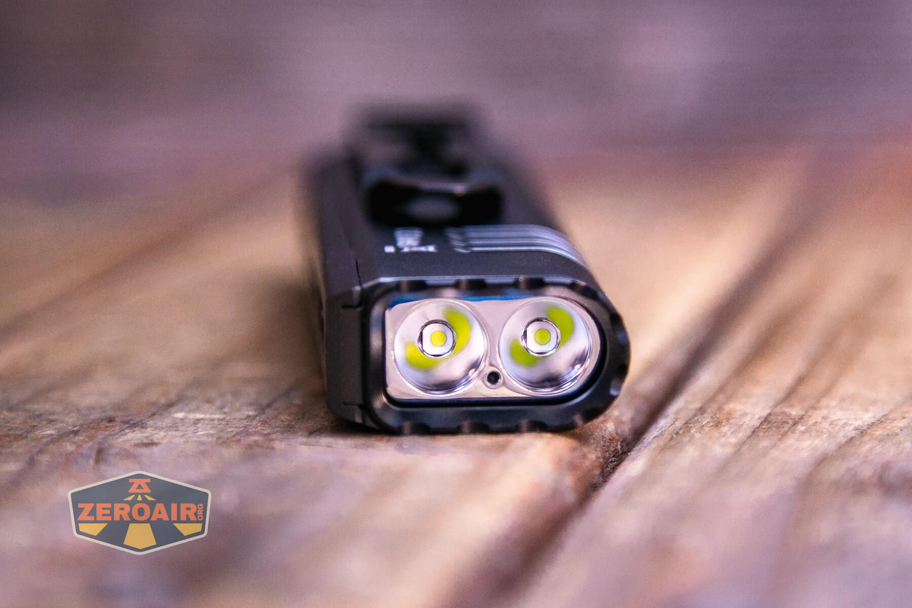 A close-up of a black dual-LED flashlight resting on a wooden surface, with the flashlights two LED bulbs facing the camera. A ZeroAir logo is visible in the bottom left corner.