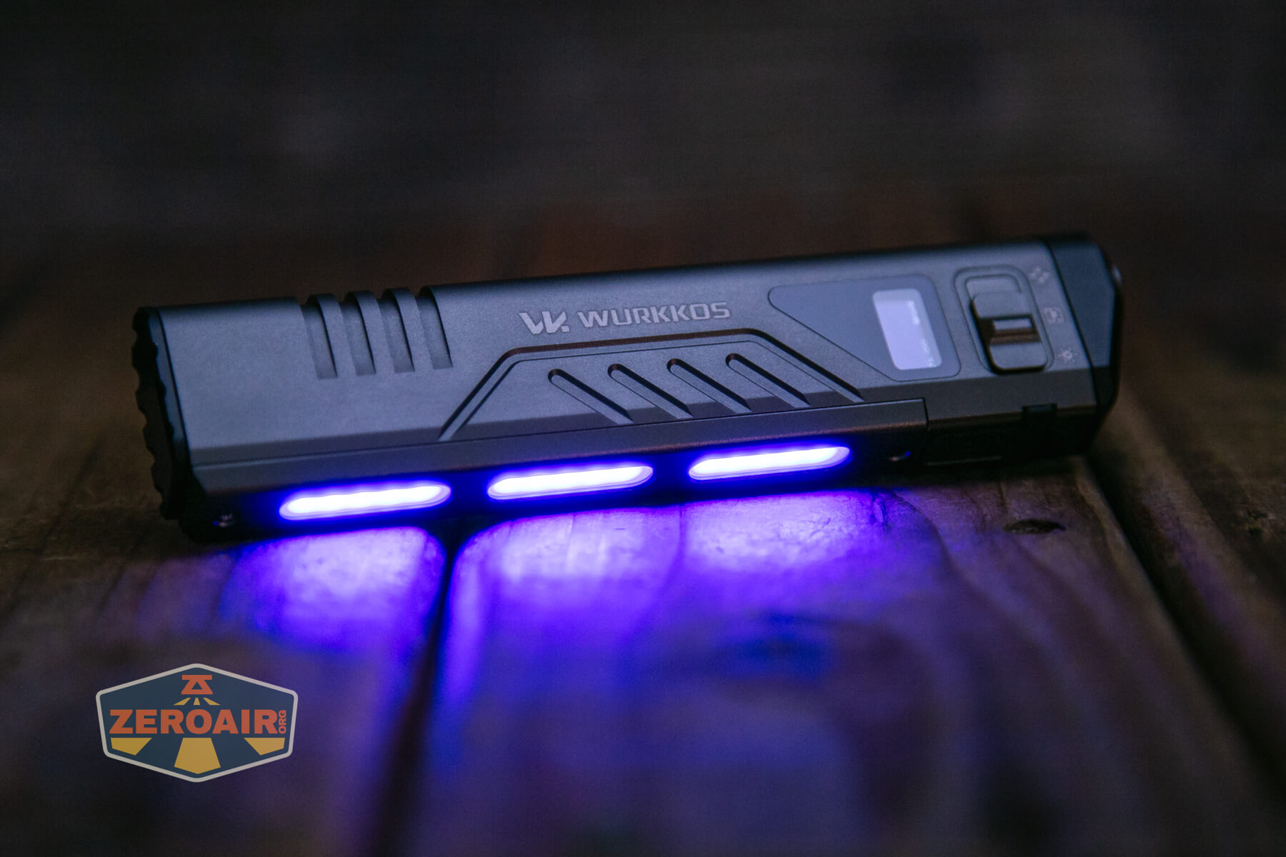 A black Wurkkos-branded flashlight lies on a wooden surface, emitting blue UV light from three bars along its side. The ZeroAir logo is visible in the corner.