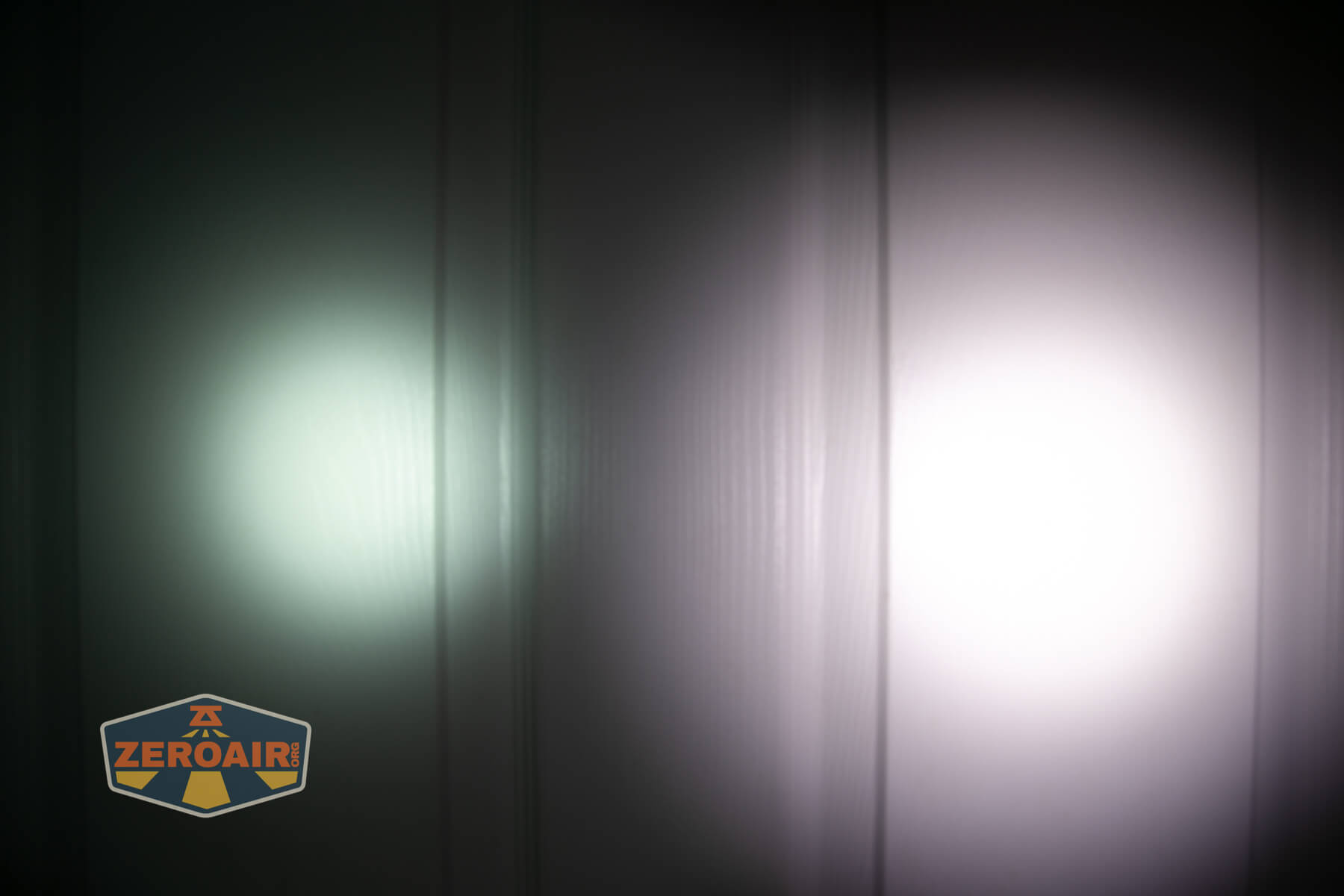 Two flashlight beams shine on a vertical, ribbed white surface; the left beam is dimmer with a green tint, while the right beam is brighter and white. A ZeroAir logo is visible in the lower-left corner.