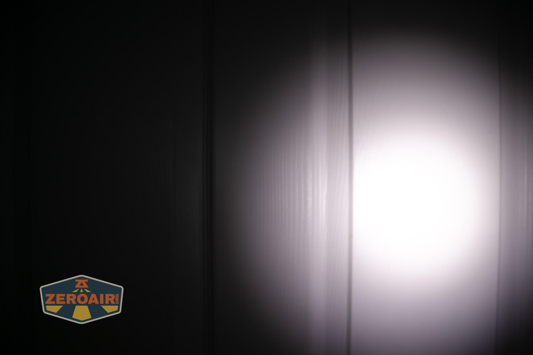 A bright circular beam of light shines on a vertically paneled wall in a dark room. The ZeroAir logo is visible in the bottom left corner.