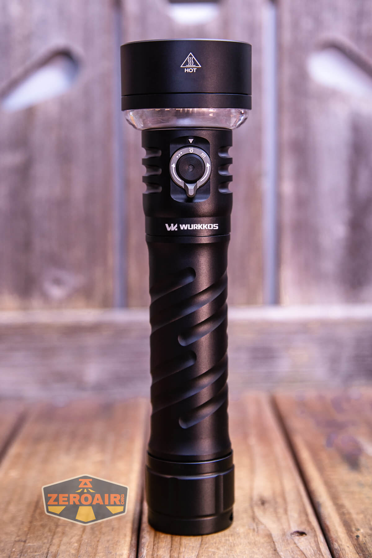 A black Wurkkos flashlight stands upright on a wooden surface with a blurred wood background. A round button and arrow indicator are visible on the flashlights body. A ZeroAir logo is in the lower left corner.