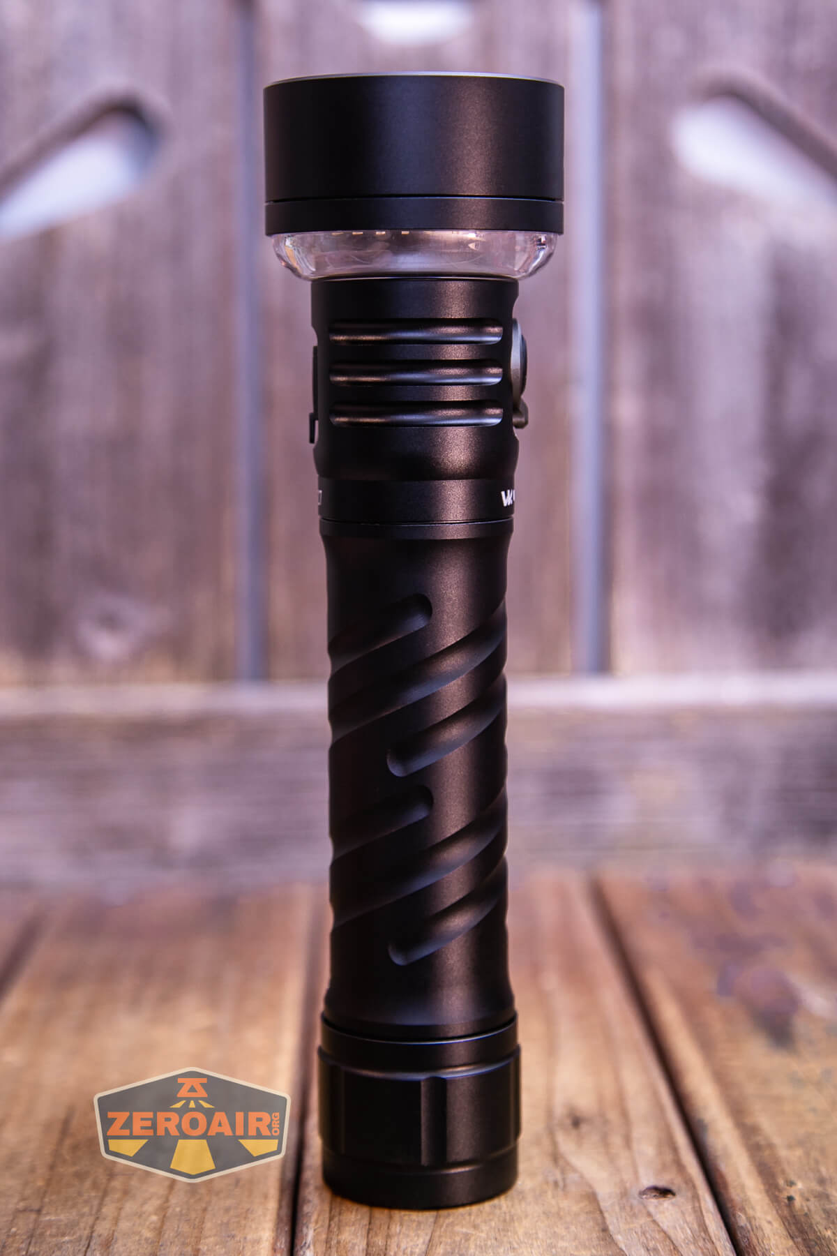 A black flashlight with a spiral design on its handle stands upright on a wooden surface. The background is wooden with a blurry ZeroAir logo in the lower left corner.