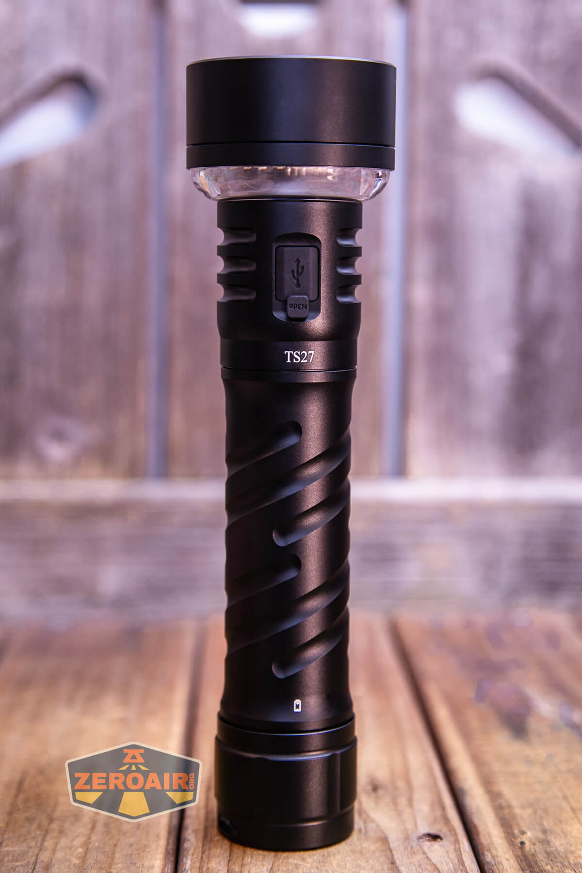 A black handheld flashlight labeled TS27 stands upright on a wooden surface with a blurred wooden background. A USB port and a button are visible on its body. The ZeroAir logo appears in the lower left corner.