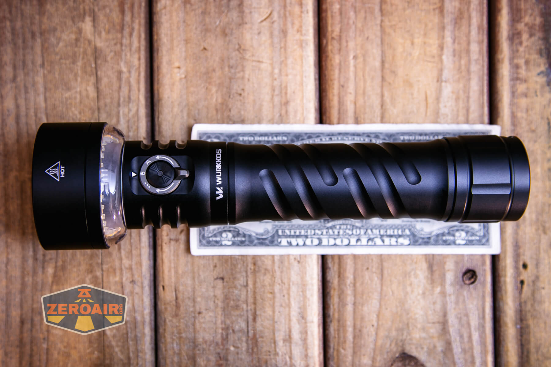 A black flashlight is placed horizontally on top of a U.S. two-dollar bill on a wooden surface. A ZEROAIR logo is visible in the bottom left corner of the image.