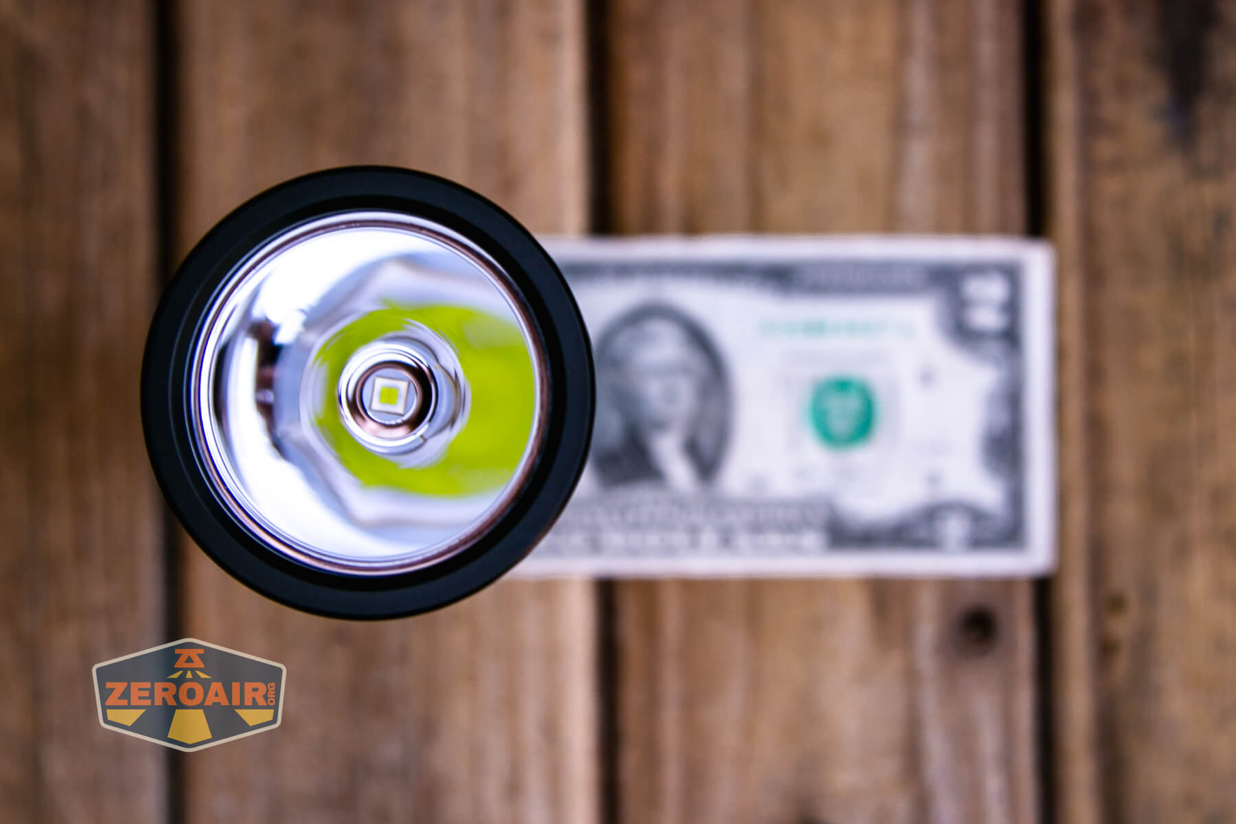 A close-up, top-down view of a flashlight lens next to a U.S. one dollar bill on a wooden surface. The ZeroAir logo appears in the bottom left corner.