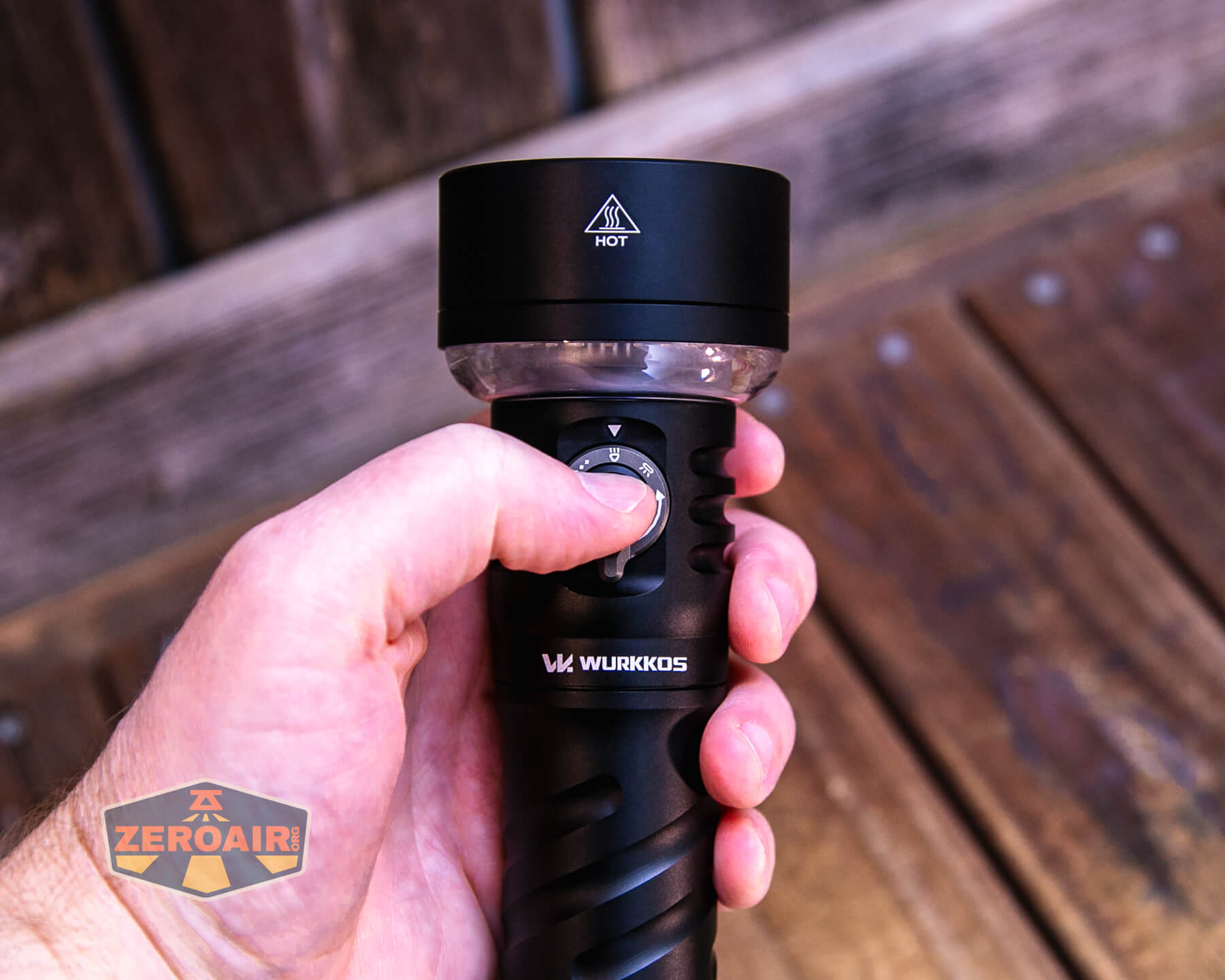 A hand holds a black Wurkkos flashlight, pressing the power button. The flashlight is labeled “HOT” on the head. The background shows wooden boards, and a Zeroair logo is visible in the lower left corner.