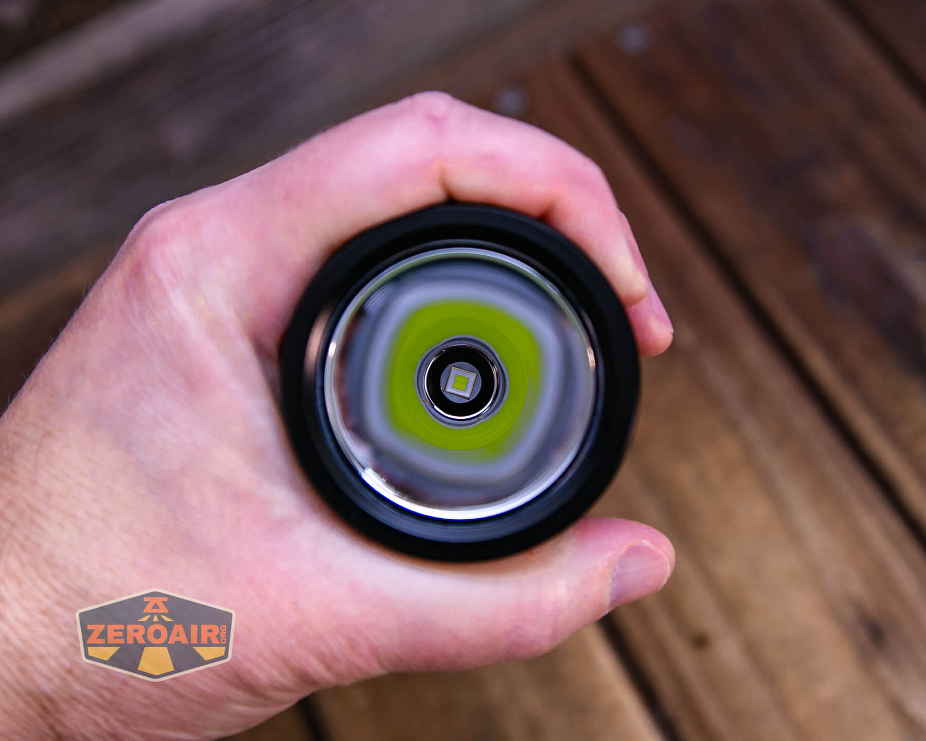 A hand holding a flashlight, viewed from the front with the lens and LED bulb facing the camera; wooden boards are visible in the background, and a ZeroAir logo is in the lower left corner.