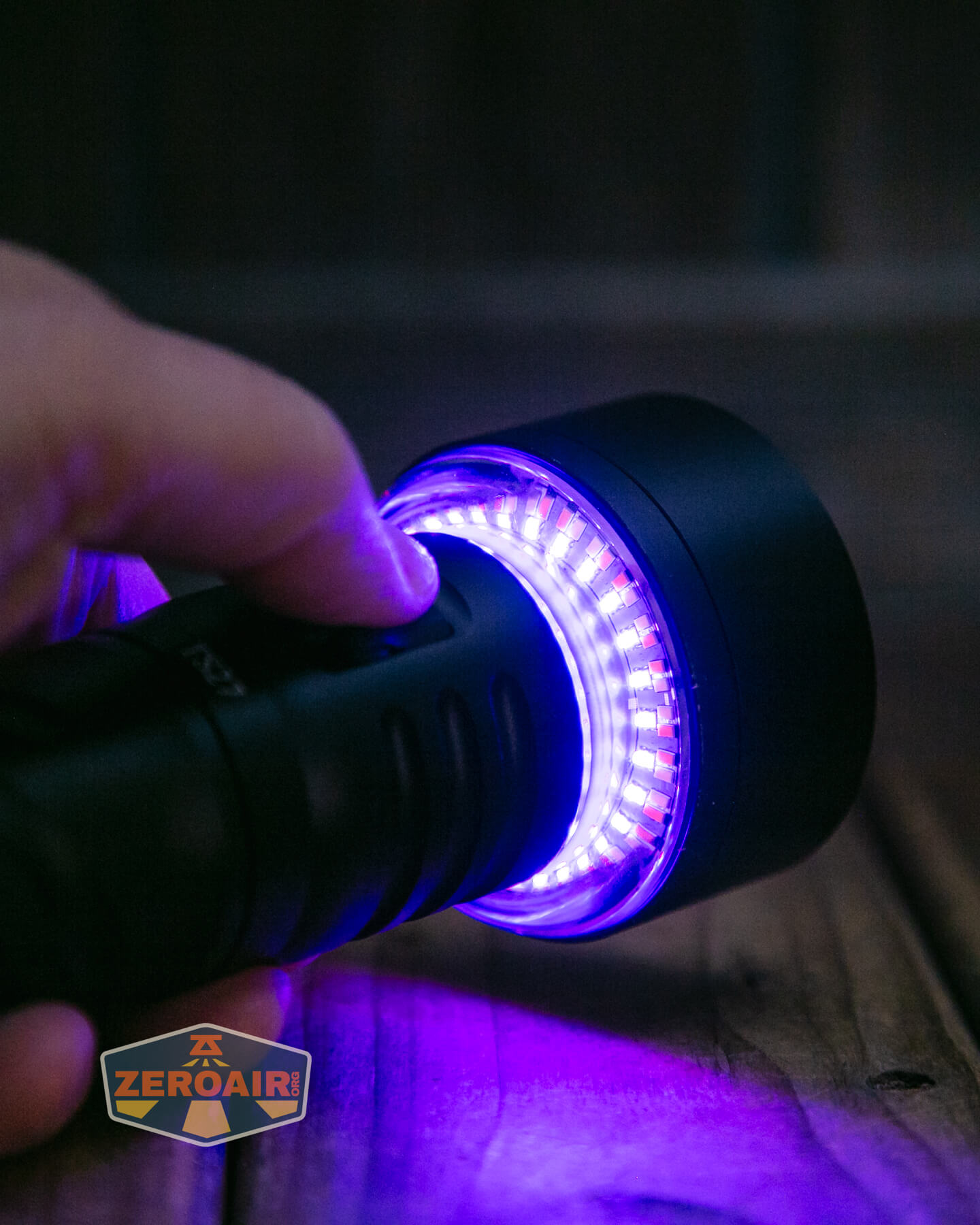 A close-up of a hand holding a black flashlight with a ring of purple LED lights glowing around the lens, positioned on a wooden surface. The ZeroAir logo appears in the bottom left corner.