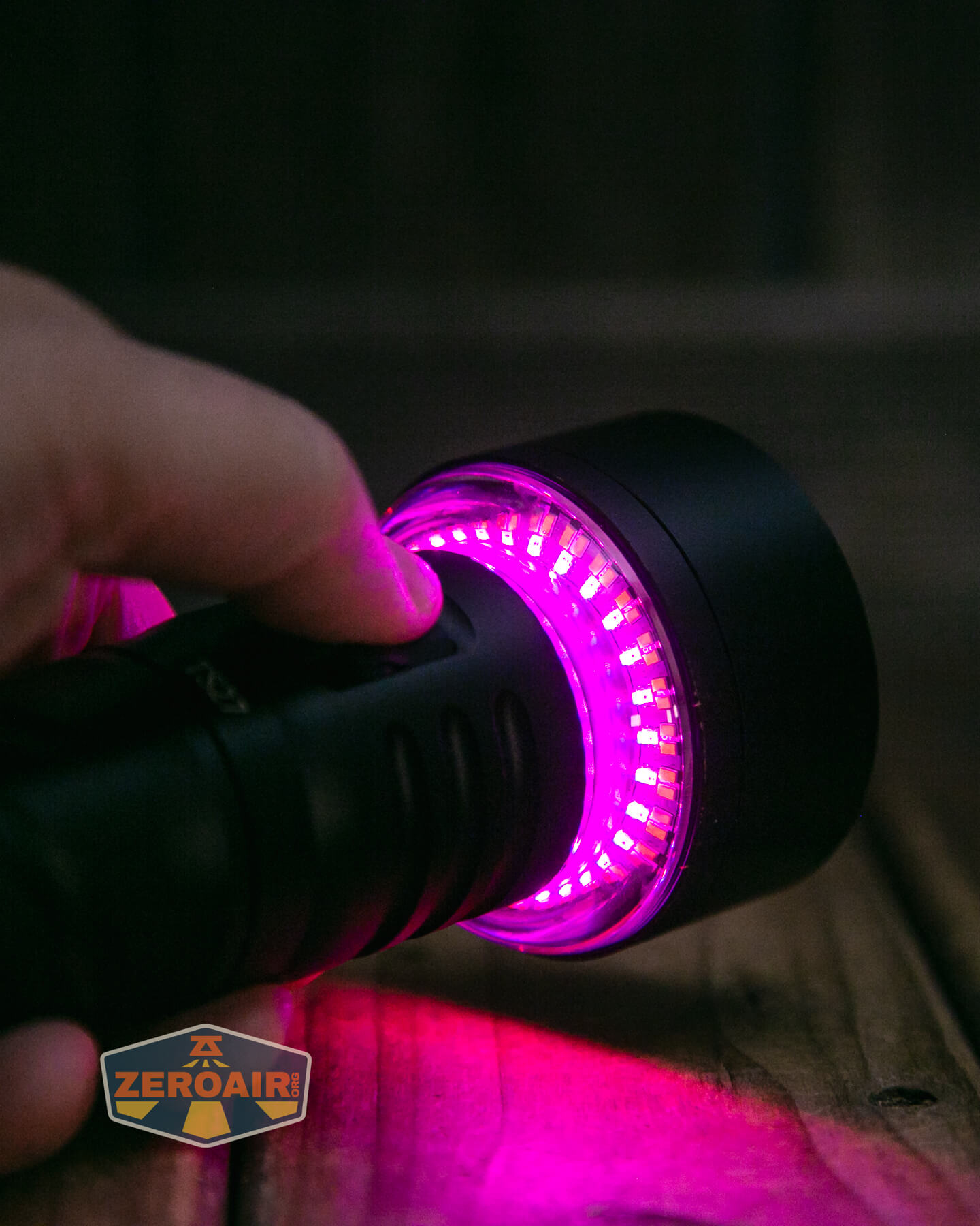 A hand holding a black flashlight emitting a bright purple light onto a wooden surface. The flashlight features multiple small LED bulbs around the lens. A patch with the text “ZEROAIR!” is in the lower left corner.