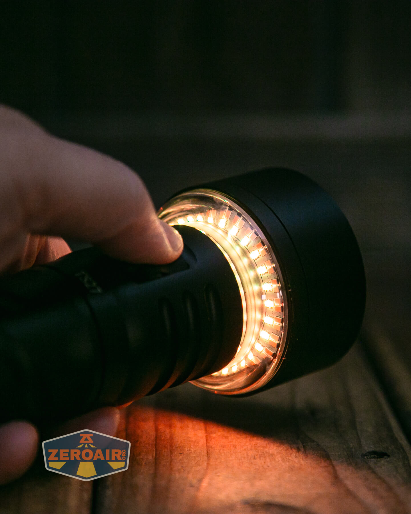 A hand holds a black flashlight with orange and white LEDs lit around the lens, casting light on a wooden surface. A ZeroAir watermark is visible in the bottom left corner.