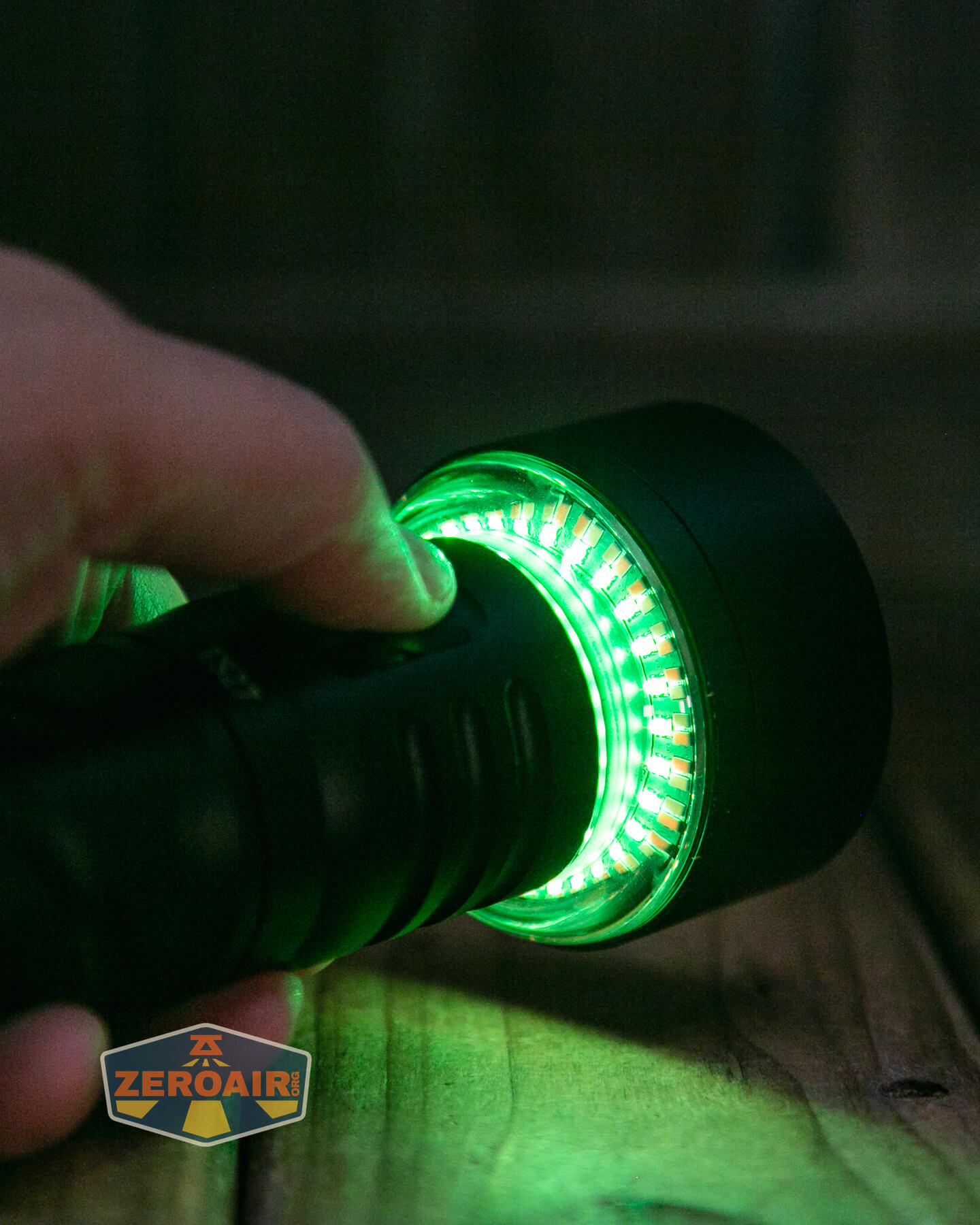 A hand holds a black flashlight with a ring of bright green LED lights illuminating around the lens, shining onto a wooden surface. A ZEROAIR logo is visible in the corner of the image.