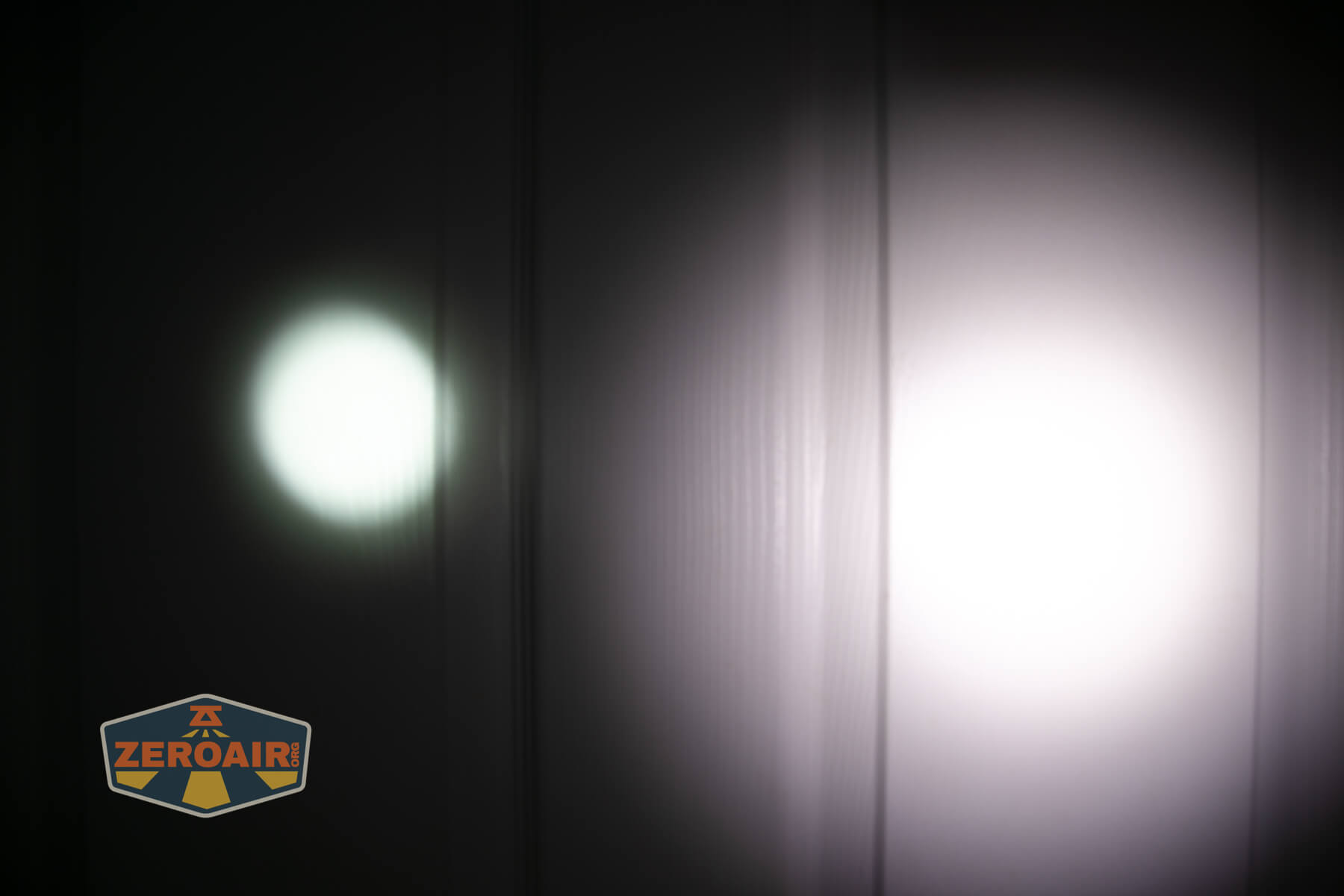Two flashlight beams are projected onto a flat, white vertical surface, with one beam on the left being smaller and dimmer, and the other on the right being larger and brighter. A ZEROAIR logo is in the bottom left corner.