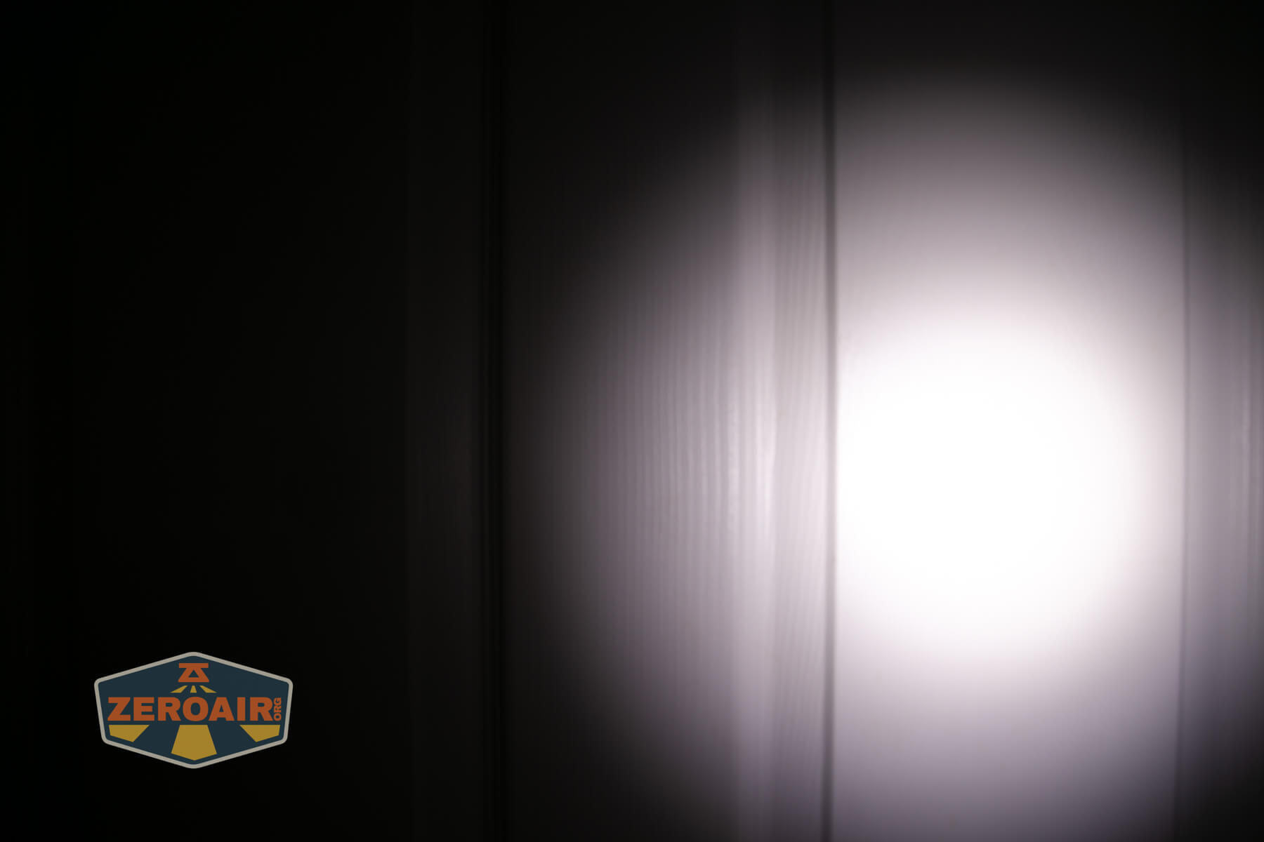 A bright white light is shining on a white vertical paneled surface, creating a circular beam effect. The rest of the image is dark. A ZeroAir logo is visible in the bottom left corner.